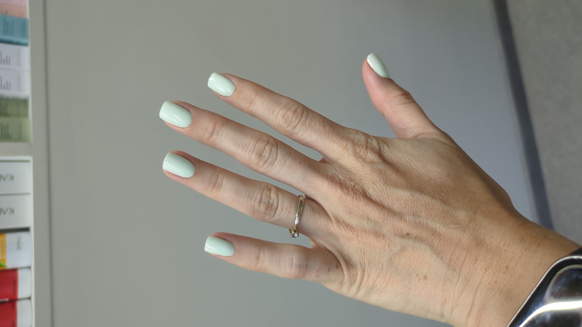 A person's left hand is displayed against a neutral gray background. The hand has a light skin tone and shows visible veins and some age spots. A silver ring with a small, clear stone is worn on the ring finger. All fingernails are painted a light, pastel green color and appear to be of medium length with a rounded shape. The tips of the fingers are visible, with the thumb positioned slightly below the other fingers. In the background, a white shelf is visible at the top, holding several product bottles or boxes, some of which have labels with text. The lower portion of the image shows a gray wall and a darker gray baseboard. This image was taken in Chișinău, Moldova.