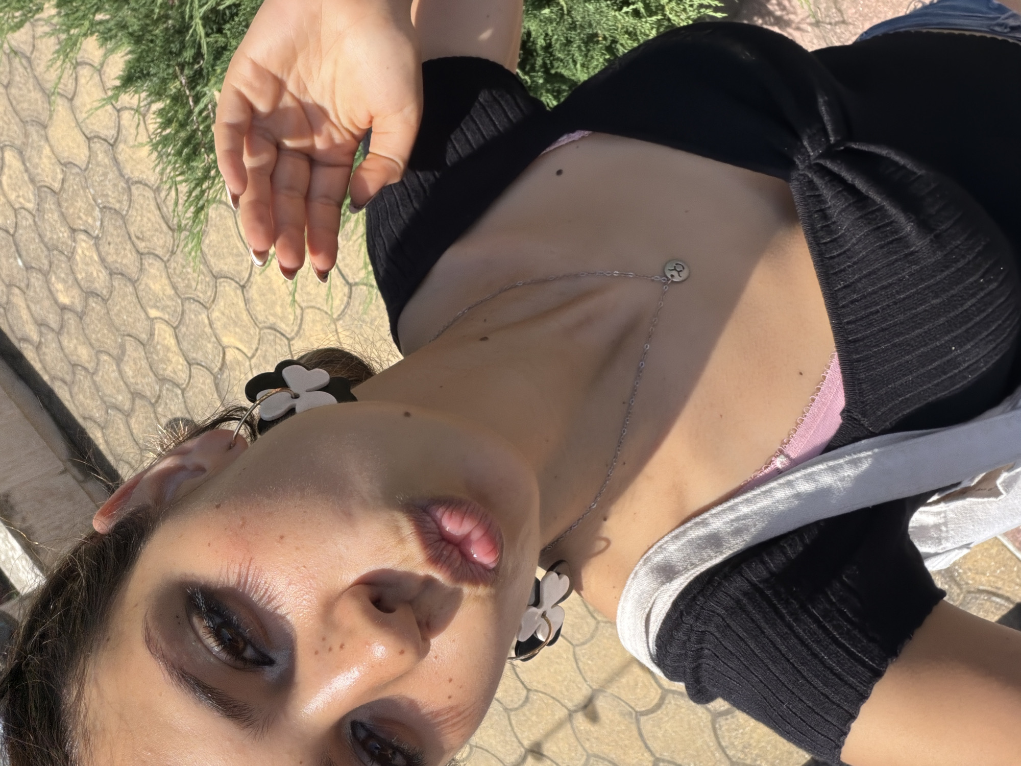 This is a vertical, close-up, selfie-style photograph of a young woman outdoors on a sunny day. She is wearing a black, ribbed top with a light pink bra underneath and a silver necklace with a circular pendant. She has dark eye makeup and is wearing large, white and black flower-shaped earrings. Her lips are pursed, and she has freckles on her face. Her right hand, with dark-tipped fingernails, is visible in the upper left quadrant of the image. The background shows a paved area with a textured pattern, and some green foliage from a conifer tree. The lighting suggests it is daytime, likely mid-morning or afternoon, given the strong shadows. The setting appears to be a public outdoor space, possibly a park or a walkway, in Lozenets, Bulgaria. No visible text is present in the image.