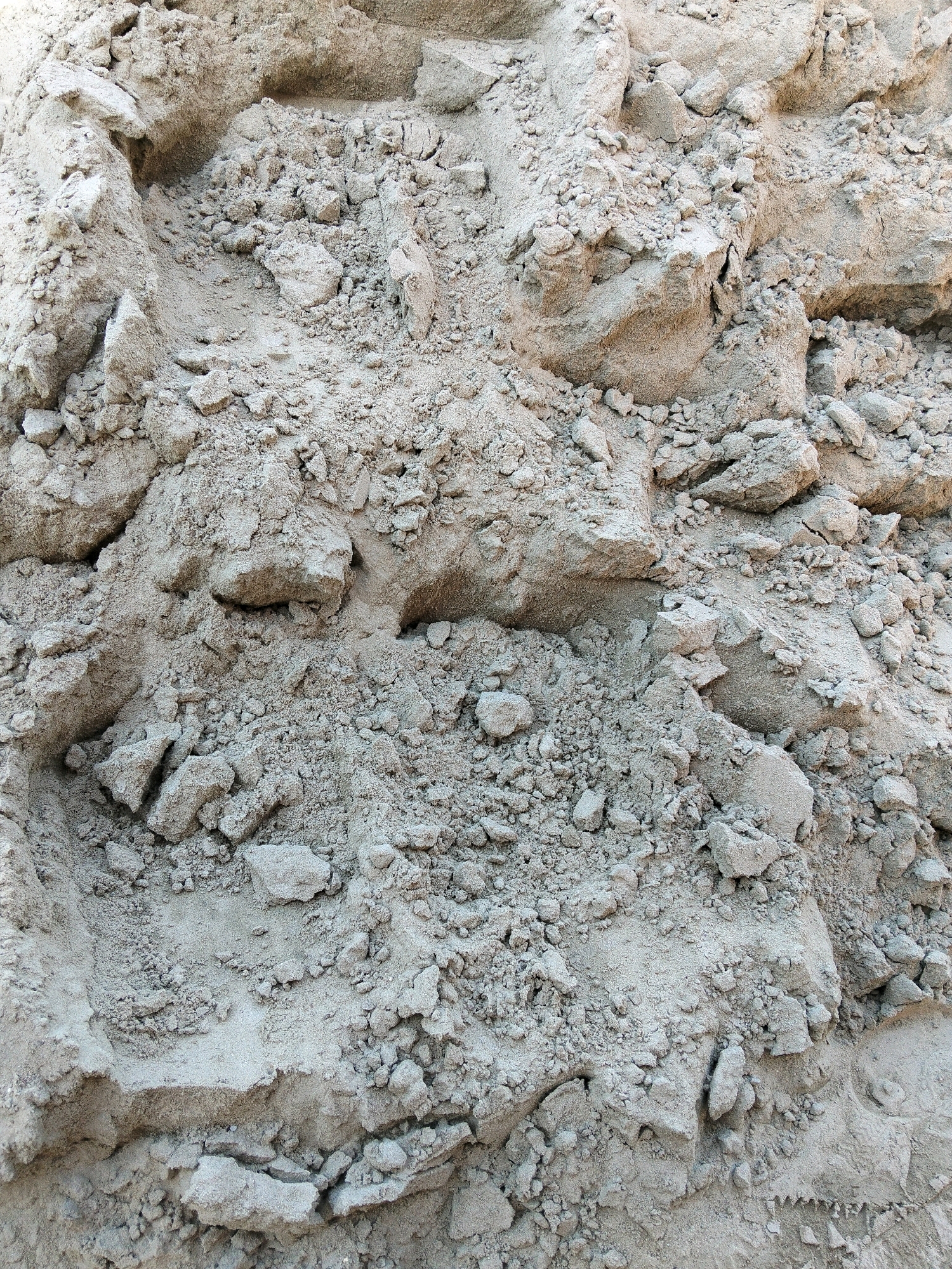 The image is a close-up, top-down view of a pile of dry, light gray sand. The sand is clumped and uneven, with visible small pebbles and larger, angular chunks of compressed sand. The texture is rough and granular. There are no discernible objects, people, or actions depicted in the image. The lighting suggests an outdoor environment, possibly under natural daylight, casting subtle shadows that highlight the uneven surface of the sand. The scene does not provide any specific details about its location in the city of Omsk, Russia, beyond being a pile of sand.