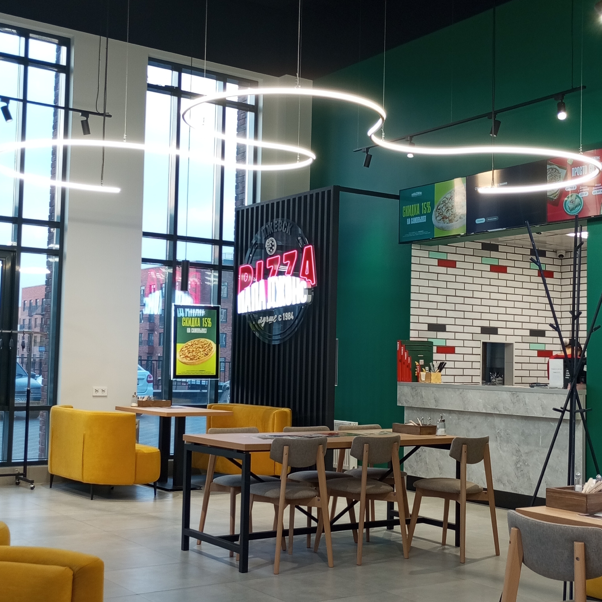 The image depicts the interior of a pizza restaurant in Izhevsk, Russia. The restaurant features a modern design with a prominent neon sign that reads "PIZZA ПАПА ДЖОНС" (Papa John's Pizza) in Cyrillic letters. The walls are painted a dark green, and the decor includes minimalist furniture, with bright yellow upholstered benches and light wood tables and chairs. Large windows offer a view of a city street with brick buildings outside. Circular LED light fixtures hang from the ceiling, casting a bright glow over the dining area. The counter area is visible in the background, with a brick-tiled wall and a marble countertop. Promotional posters displaying pizza and discounts are mounted on the wall above the counter. The scene is clean and well-lit, suggesting a typical commercial establishment. There are no visible people in the frame.