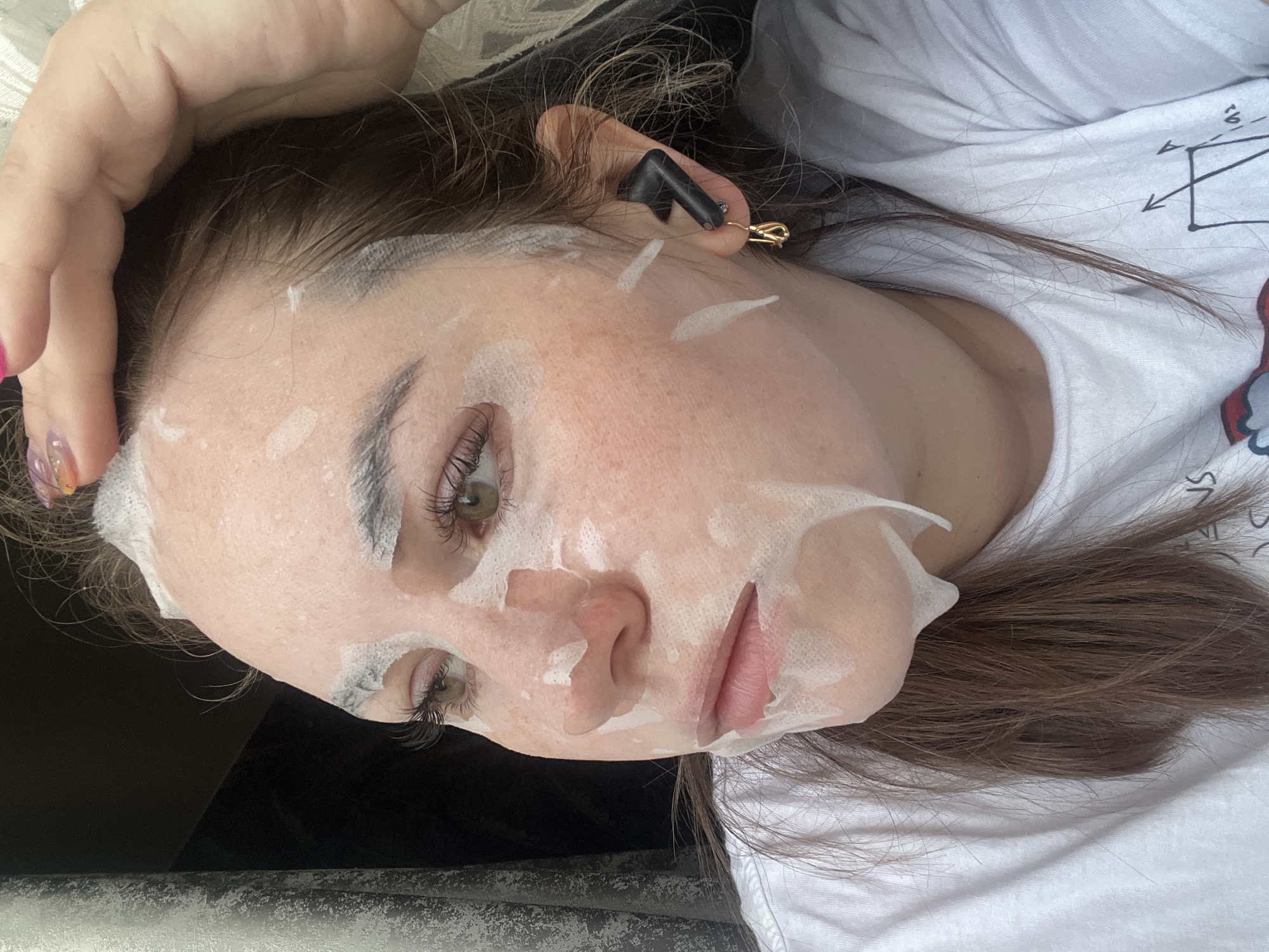 The image displays a close-up, overhead view of a person lying down, seemingly in a relaxed state. The subject is a female with fair skin, dark brown hair, and light green or hazel eyes. Her face is covered with a white sheet mask, which has cutouts for her eyes, nostrils, and mouth. Liquid or serum from the mask is visible on her skin. Her eyelashes appear long and dark, possibly enhanced with mascara or extensions, and her eyebrows are well-defined. Her lips are a natural pink.
In her right ear (left side of the image), she wears a black wireless earbud, and a small, gold-colored safety-pin-like clip or earring is also visible near her earlobe. Her left hand is partially visible near the top of her head, showing manicured nails with a colorful, possibly gradient design. She is wearing a white t-shirt that features faint black graphics, including a partial diagram with arrows, resembling a scientific or mathematical illustration. Her long dark hair is spread out behind her.
The background directly behind her head is dark, while the surface she is lying on appears to be a lighter, possibly white or light grey material. The overall scene suggests a quiet, indoor setting, likely a self-care or relaxation moment. The person's expression is calm and neutral.