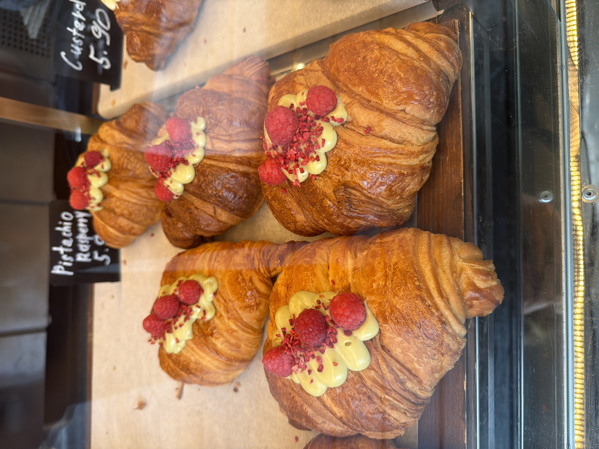 The image shows a display of croissants in a bakery, likely in London, United Kingdom. The croissants are golden brown and appear to be freshly baked. Each croissant is decorated with a dollop of pale yellow cream and topped with three fresh raspberries and some small pieces of red dried raspberry. A small black sign is visible to the left, displaying the words "Pistachio Raspberry 5.90", indicating the flavor and price. Another black sign displays the words "Custard 5.90". The pastries are behind a glass panel, suggesting they are in a display case. The background is blurred, but it appears to be a bakery or cafe setting. The environment suggests a high-quality pastry shop with gourmet or specialty items.