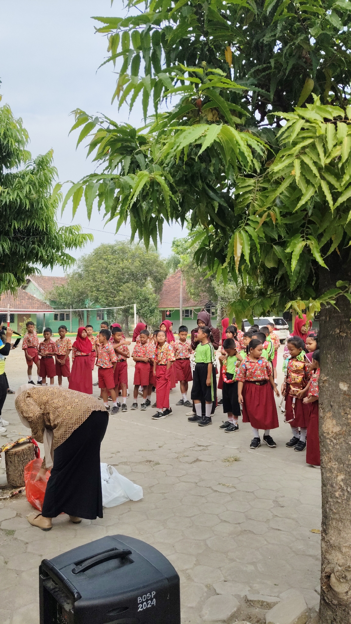 An outdoor group photo shows approximately thirty Indonesian schoolchildren, likely elementary age, dressed in red and white school uniforms, gathered in a courtyard. Some of the children wear headscarves. A woman in a patterned headscarf and a long dark skirt is bending down, interacting with items on the ground. In the foreground, a black speaker with "BOSP 2024" written on it is visible. In the background, there are low-rise school buildings with red roofs, a volleyball net, and trees. The ground is paved with hexagonal stones. The image appears to be taken in Banjarsari, Indonesia.