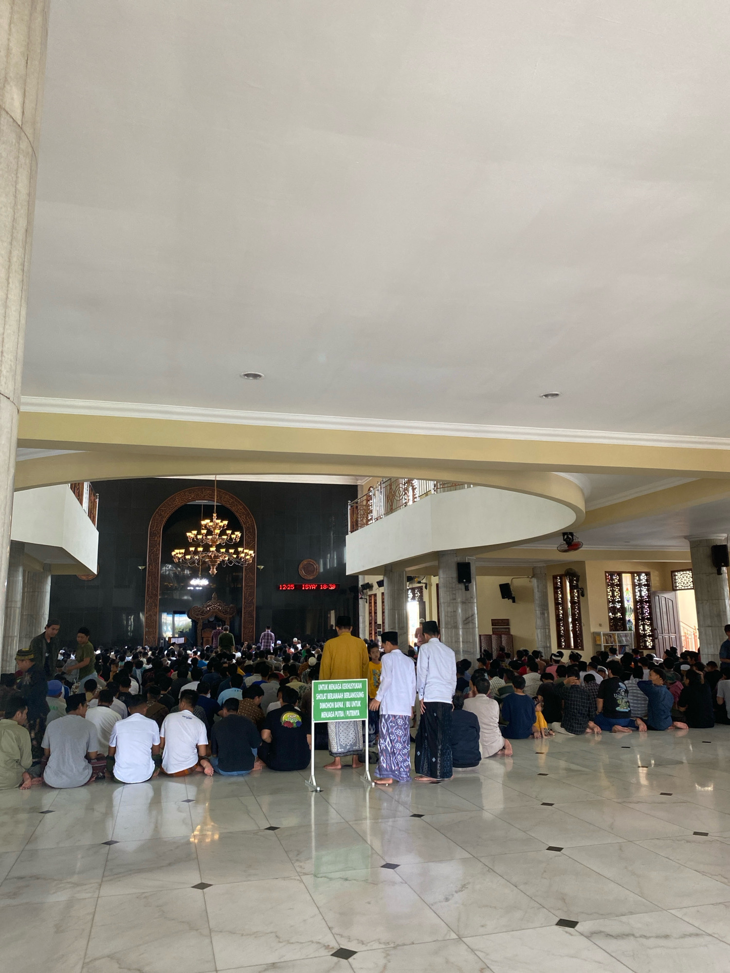The image depicts the interior of a mosque in Kediri City, Indonesia, where a large gathering of men and boys are seated on the floor, facing towards the front of the mosque, likely for prayer. The mosque has a grand architectural style with high ceilings, large pillars, and an ornate chandelier. The floor is made of polished marble tiles. In the background, there is a large arched doorway with intricate carvings, and a digital clock displaying "12:25 ISYA' 18:39". A green sign with Indonesian text is visible in the foreground. The lighting appears to be a mix of natural light from windows and artificial light from the chandelier and ceiling fixtures. The atmosphere seems solemn and respectful, with individuals dressed in modest Islamic attire.