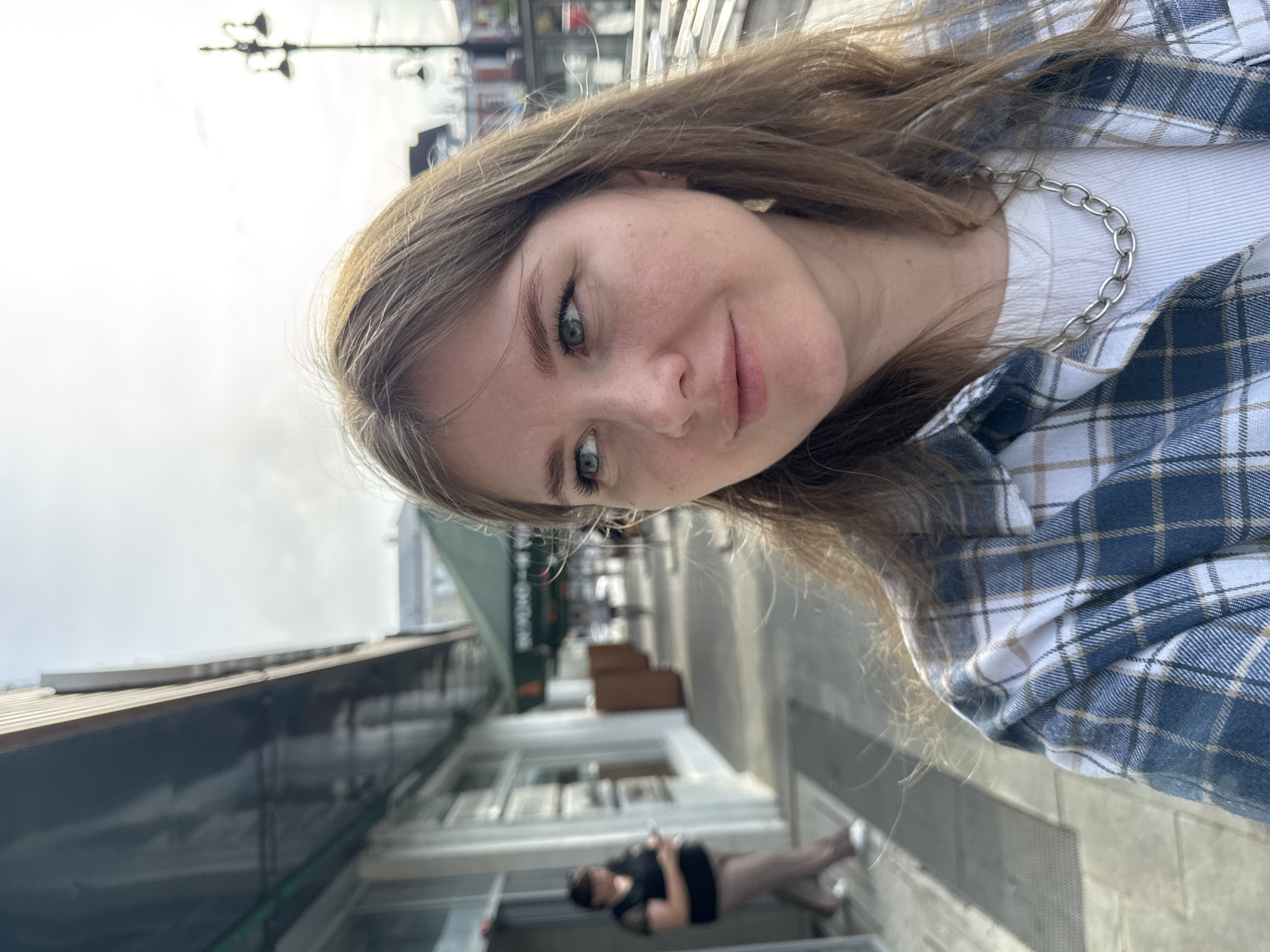 A young woman with light brown hair and blue eyes is taking a selfie. She is wearing a white top with a silver chain and a blue and white plaid shirt. In the background, there is a building and a sidewalk. A woman in black tights and black shirt, is sitting on a window ledge in the background. Based on architectural style, this is most likely located in the city of Perm, Russia. The environment appears to be overcast, with the sky visible in the background. The photo is taken at an angle, with the woman tilting her head slightly.