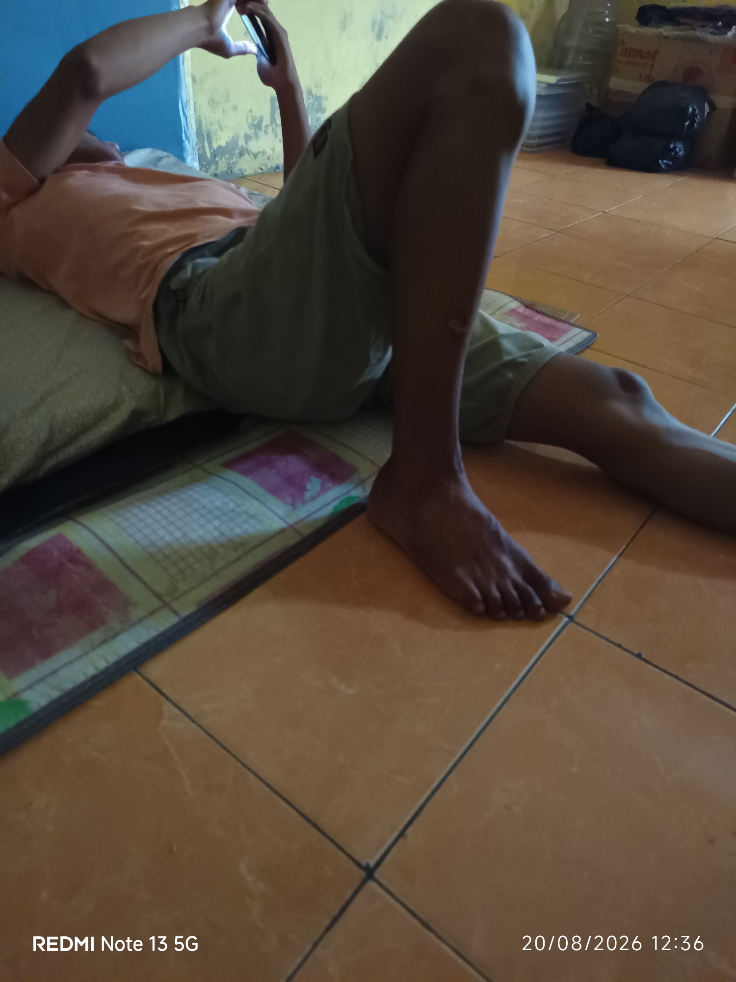 A person is lying on their back on a floor, using a mobile phone. The individual is wearing a light orange t-shirt and light-colored shorts. Their legs are bent, and their feet are visible. The person is holding the phone in their hands, seemingly engaged with its screen. The floor appears to be tiled in an orange-brown color. A patterned mat or cushion is visible beneath the person's body, and part of a wall painted blue and yellow is in the background. There are also some items in the background, including what appear to be boxes and black plastic bags. The timestamp in the lower right corner indicates the image was captured on August 20, 2026, at 12:36 PM, and the lower left corner shows "REDMI Note 13 5G", suggesting it was taken with a Redmi Note 13 5G phone. The location is identified as Talaga Sari, Indonesia.