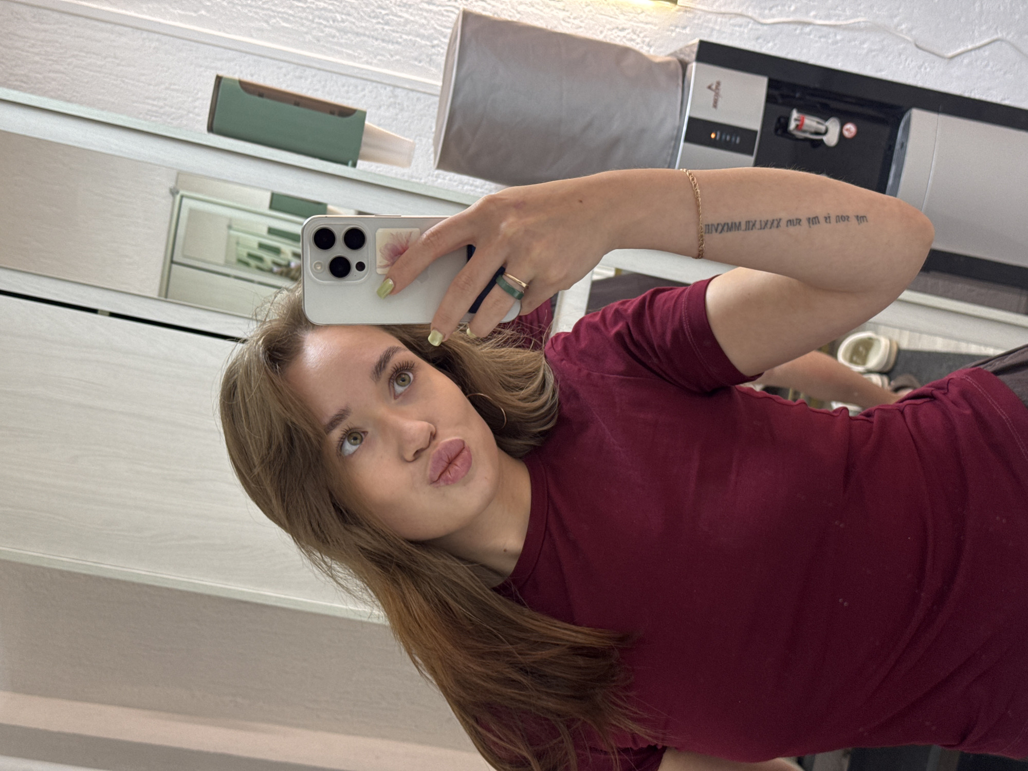 A young woman with blonde-brown hair and green eyes is taking a mirror selfie indoors. She is wearing a maroon t-shirt and has light green nail polish. She is holding a silver iPhone in her right hand, which is adorned with two rings and a gold bracelet. A tattoo is visible on her left forearm. The background includes a mirror reflecting another room, a dispenser for paper towels, and a water cooler. The scene appears to be in a public facility or an office building, possibly in the city of Chaikovsky, Russia.