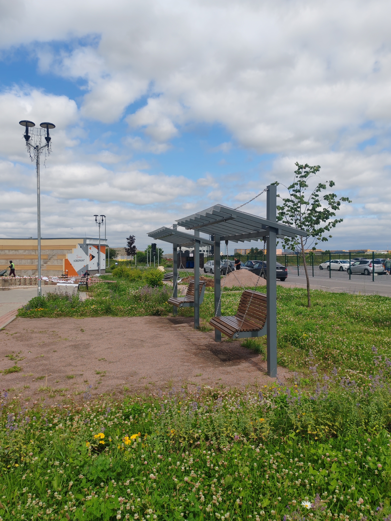 The image depicts a public park area in Shushary, Russia, on a cloudy day. In the foreground, a modern covered seating area with two wooden swings is situated on a gravel-covered ground. The swings are suspended by chains from a gray metal frame with a translucent roof. Adjacent to the seating area, a patch of lush green grass is filled with small white and purple wildflowers, and a few yellow flowers. In the middle ground, a paved pathway runs alongside the grassy area, and further back, a building with tiered seating is visible. A tall lamppost with two lights and decorative string lights stands to the left. A person is seen pushing a wheelbarrow on the pathway. In the background, a parking lot with several cars is present, and beyond that, a row of buildings. A young tree with sparse leaves grows near the covered seating area. The sky is filled with white cumulus clouds against a blue background.