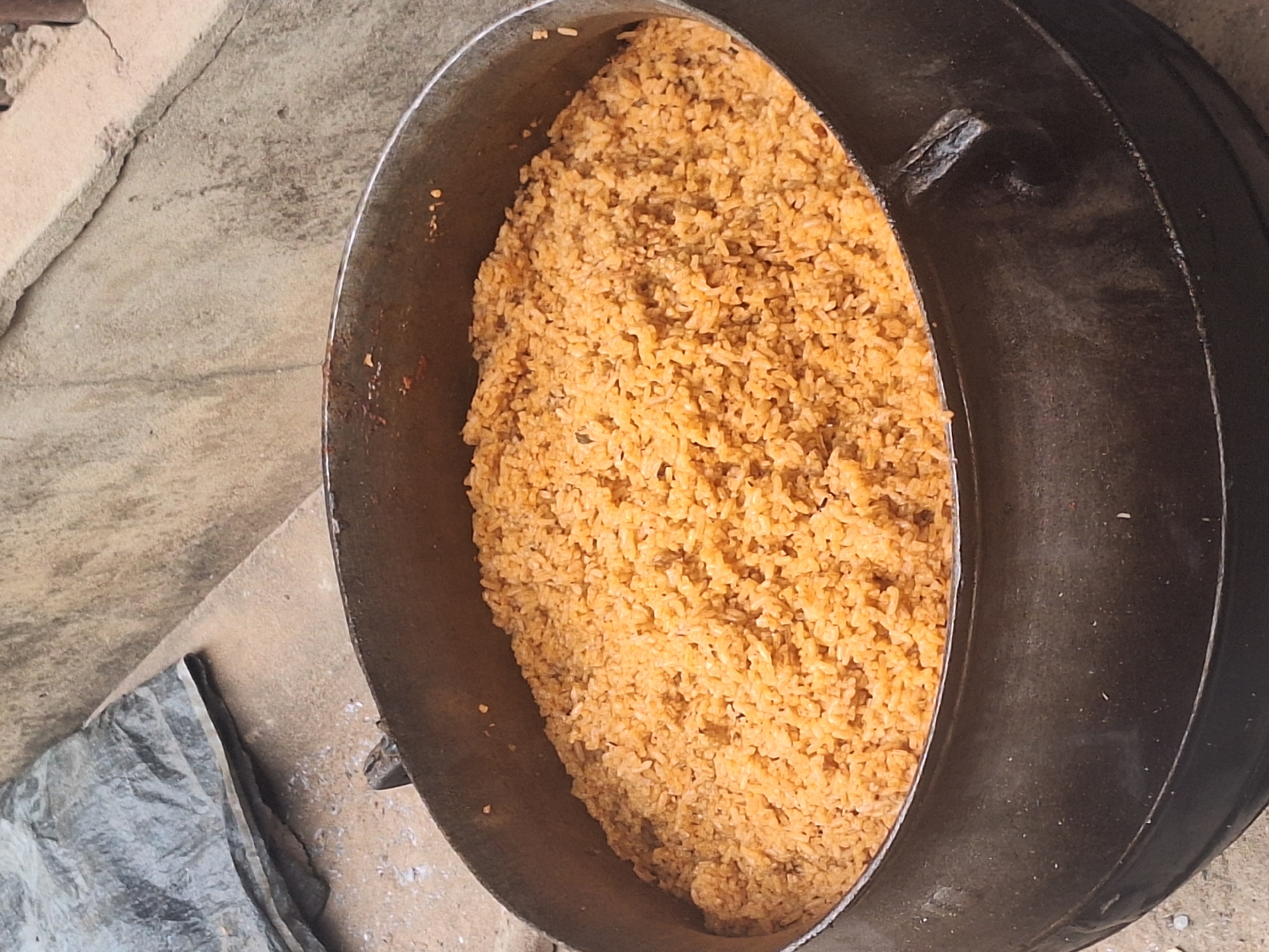A top-down, high-angle shot shows a large, well-worn metal pot filled with orange rice. The pot sits on a concrete surface, likely outdoors, with a piece of gray plastic tarp visible on the left. The location is implied to be Sabon Tasha, Nigeria, based on the prompt. No people are visible. The subject is the pot of rice.