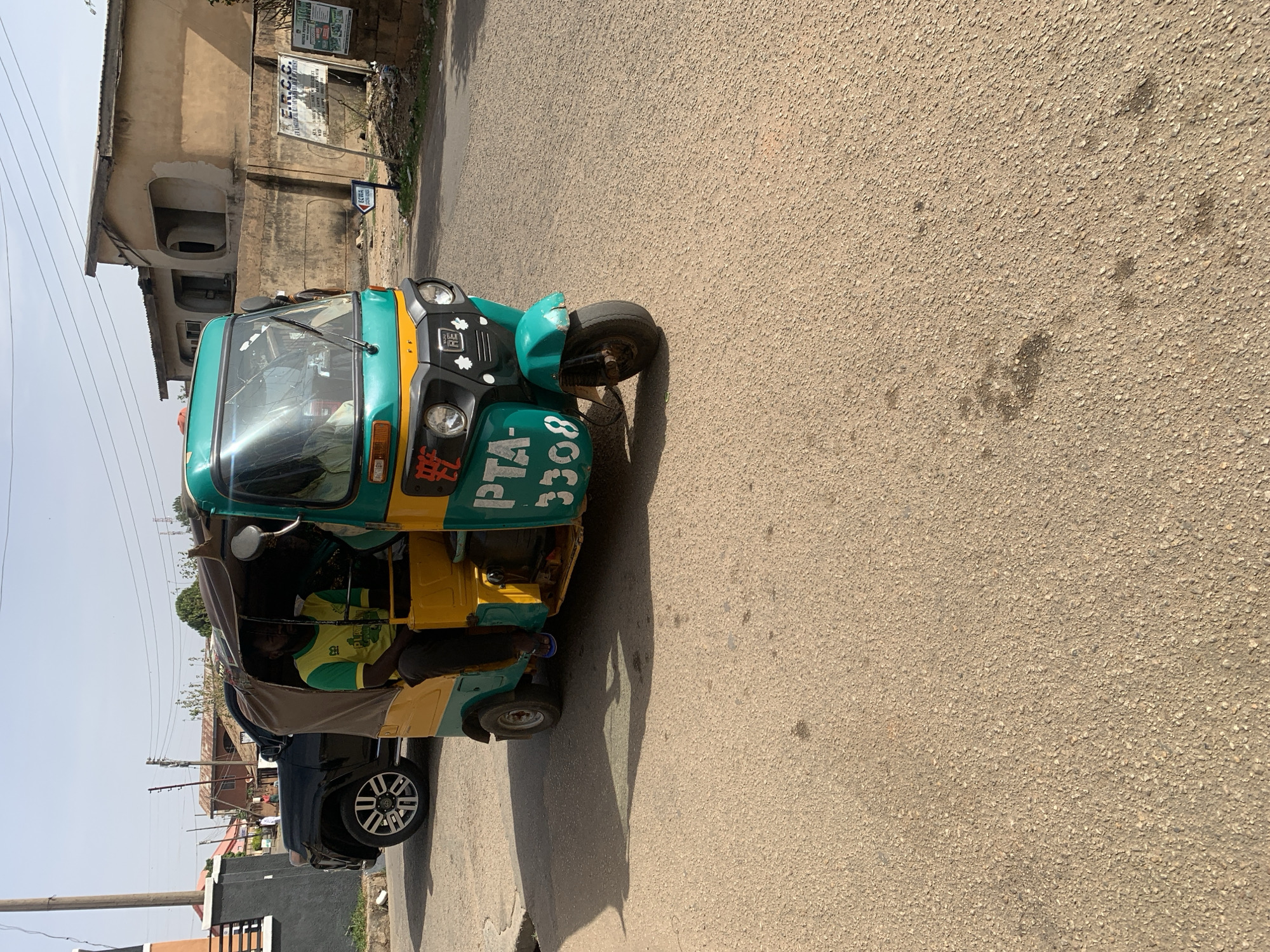 The image captures a vibrant green and yellow auto-rickshaw, commonly known as a Keke Napep, on an asphalt road in what appears to be an urban or semi-urban setting in Jos, Nigeria. Bright sunlight casts strong shadows, suggesting it is midday or early afternoon under a clear, dry sky.
Inside the auto-rickshaw, two men are visible: a driver in a green and yellow shirt at the wheel and a passenger seated behind him. The auto-rickshaw is either parked or moving slowly, with another dark-colored vehicle partially visible to its left.
In the background, a multi-story building with a light-colored, textured facade and arched window/door openings is prominent. Power lines and utility poles are also visible. A sign affixed to the building reads "F.R.C.C." with "FEDERAL ROADS OF NIGERIA" discernible below it, indicating a road project. Another smaller sign nearby displays "190923".
The auto-rickshaw bears the registration number "PTA-3308" in green lettering on a yellow background. Other visible text includes red Chinese characters on a white sticker on the front, and a white letter "R" enclosed in a square emblem on the vehicle's front grille. The asphalt road surface is coarse and shows scattered dark stains.