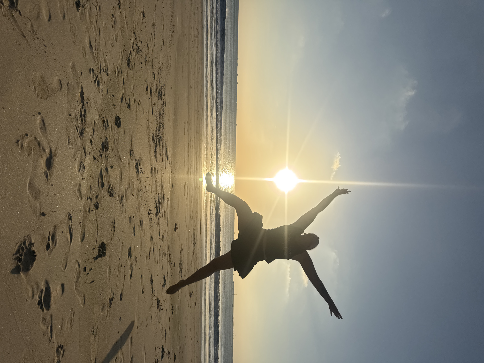 The image depicts a person silhouetted against a bright sunset on a beach. The person is captured mid-air, jumping with arms outstretched, suggesting a moment of joy or freedom. The sun is low on the horizon, casting a warm glow and creating a lens flare effect. The ocean is visible in the background with gentle waves and a few boats dotted on the water. The beach is sandy with footprints scattered across the surface. The sky is a gradient of blue and orange, with a few wispy clouds. The overall atmosphere is serene and uplifting, characteristic of a sunset scene. This image is likely taken in Kuta, Bali, Indonesia, given the characteristic wide sandy beaches, consistent waves, and popular sunset viewing spots.