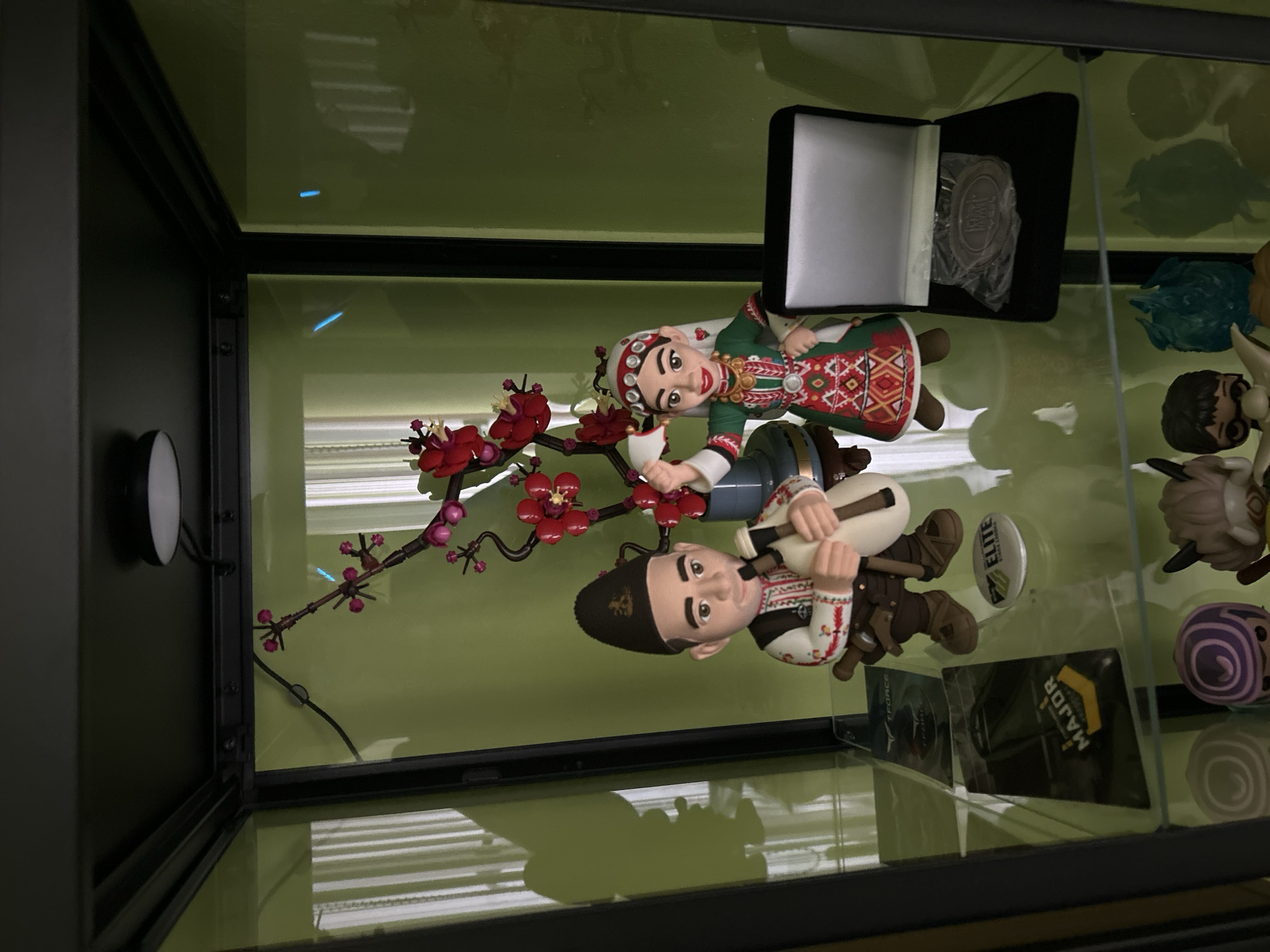 The image shows the interior of a display cabinet, likely located in a home, with several objects arranged on the shelves.
On the first shelf, there are two figurines of people in traditional Bulgarian clothing holding a decorative branch with red flowers. On the upper shelf, to the right of the figurines, there are several other collectible figurines. Also on the upper shelf, there is a black box with something white (possibly paper or fabric) inside, as well as a circular medal wrapped in plastic. Further down there are at least two cards with branding.
The location is presumed to be Plovdiv, Bulgaria, as the figurines are dressed in traditional Bulgarian clothing and the display seems to be in a domestic setting.