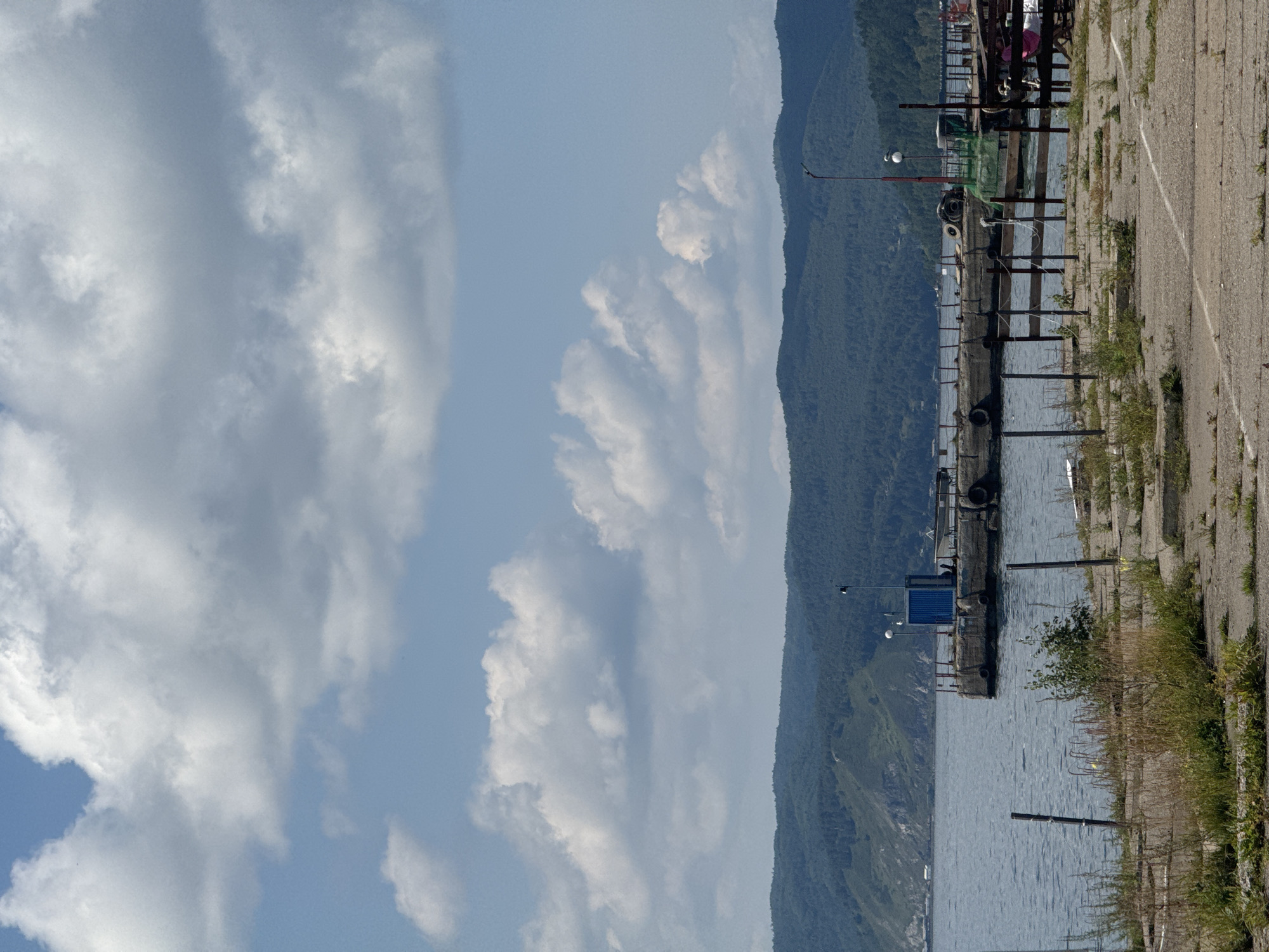 The image depicts a view of Lake Baikal near Listvyanka, Russia. In the foreground, a paved area with scattered vegetation and remnants of fencing suggests a disused or undeveloped waterfront. A dock juts out into the calm, blue water, where a small, blue structure sits. The dock is reinforced with old tires. In the background, a range of forested hills forms the horizon, with scattered buildings visible on the distant shore. The sky is partly cloudy with large, white cumulus clouds. No people are visible in the scene.