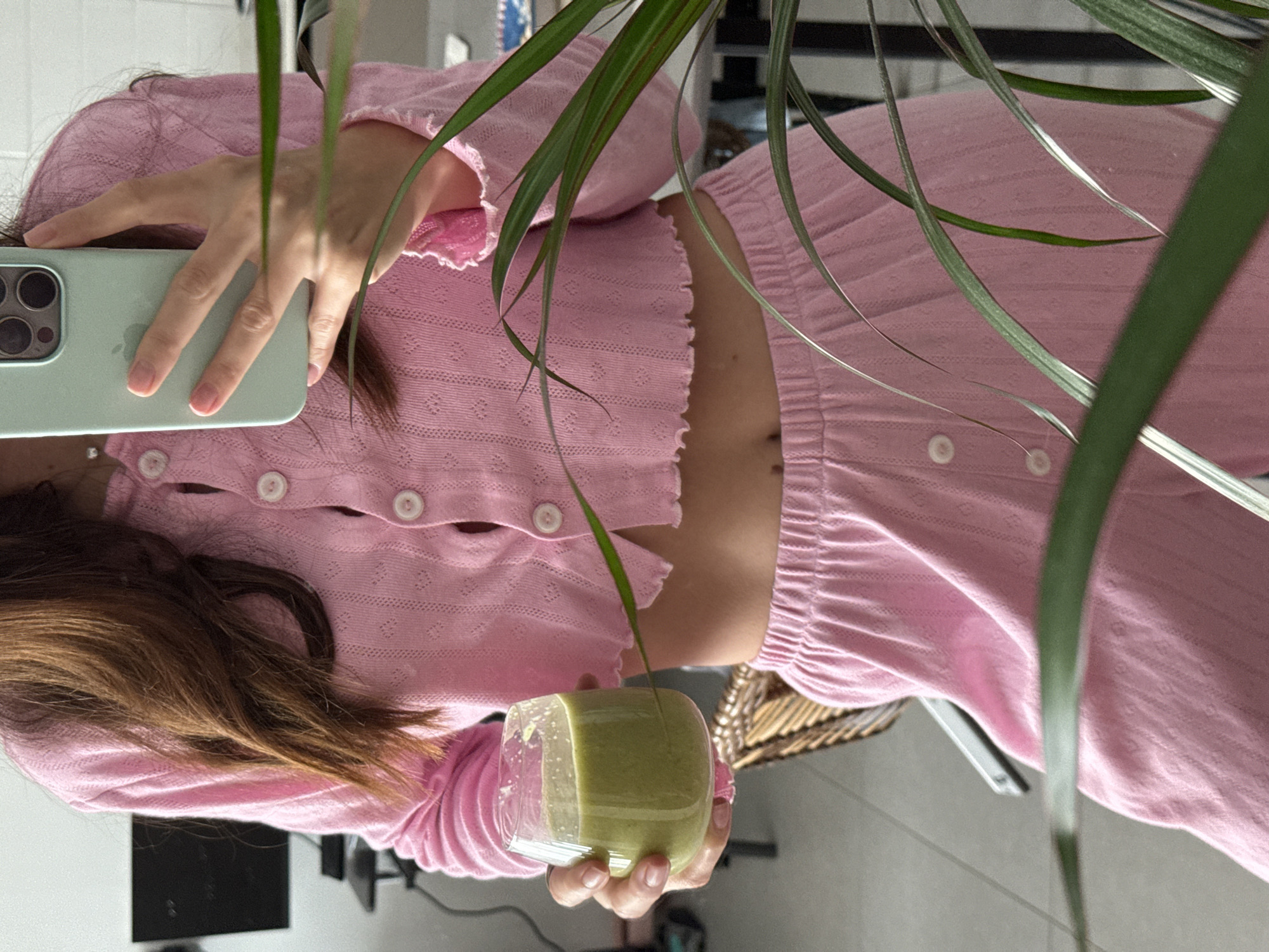 An indoor, portrait-oriented image, rotated 90 degrees counter-clockwise, depicts a person taking a mirror selfie in Ramat Gan, Israel. The subject, likely female, has long brown hair and is dressed in a matching pink two-piece ensemble. The outfit includes a cropped, long-sleeved top with a button-down front, ruffled edges, and a textured, possibly eyelet or knitted pattern. This is paired with high-waisted pink pants of a similar textured fabric, revealing a portion of the person's midriff.
In their left hand, the person holds a light green smartphone, featuring a visible camera array and an Apple logo on its case, positioned as if capturing their reflection. Their right hand holds a clear, round glass filled with a vibrant green liquid, likely a smoothie or juice.
The foreground is partially obscured by lush green plant leaves, adding a natural element to the scene. The background is blurred but suggests an interior setting, possibly a room with hints of furniture or reflective surfaces, and a woven basket-like object. The lighting indicates it is daytime. No explicit text is visible in the image apart from the Apple logo on the phone.