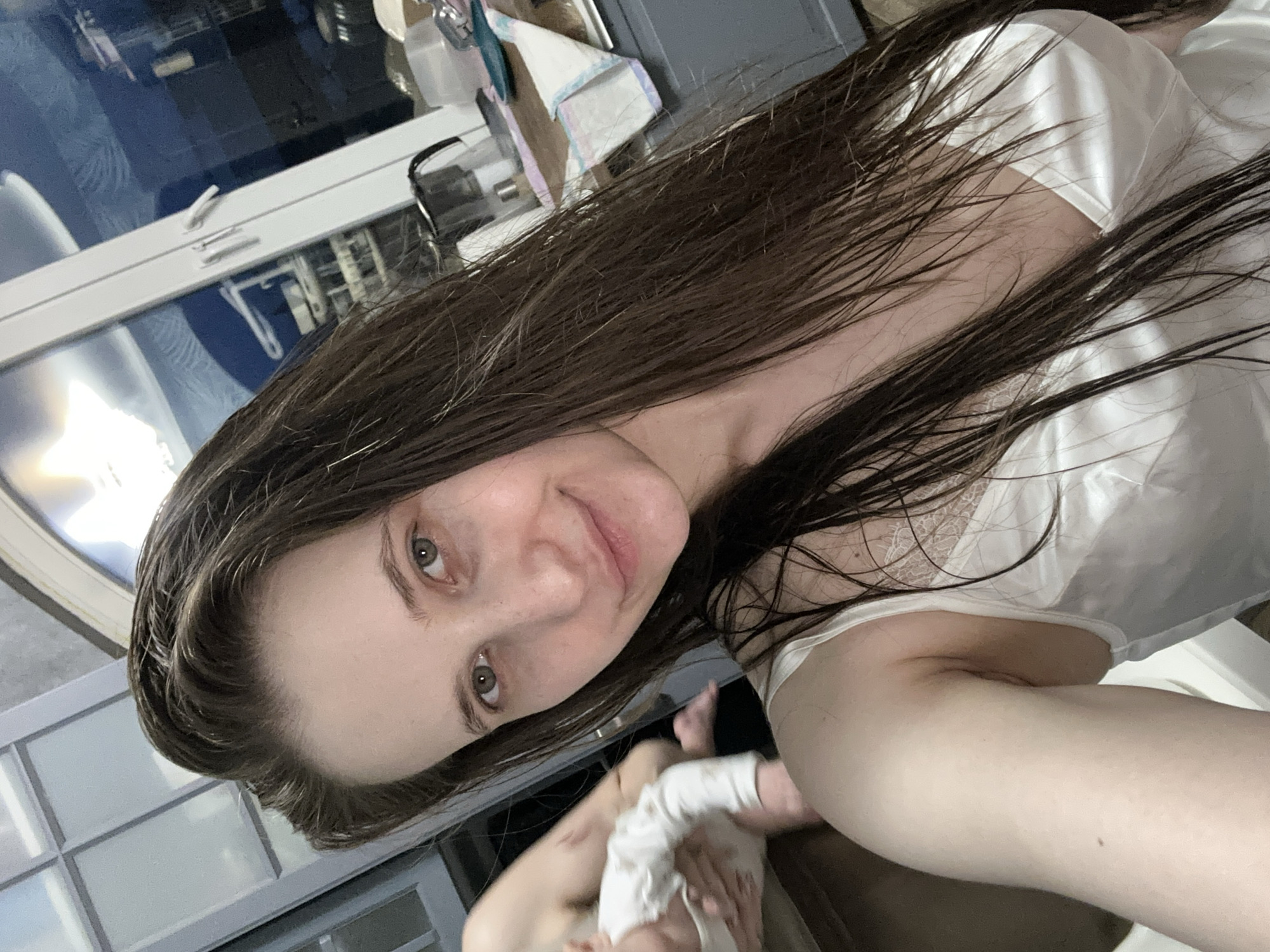 A selfie depicts a woman with wet, brown hair and green eyes. She is smiling slightly at the camera and is wearing a white top with lace detailing. She is holding a baby in her arms, who is wearing white clothing. The background shows a window with a reflection of what appears to be an outdoor scene. The location appears to be indoors in the city of Pokrov, Russia.
