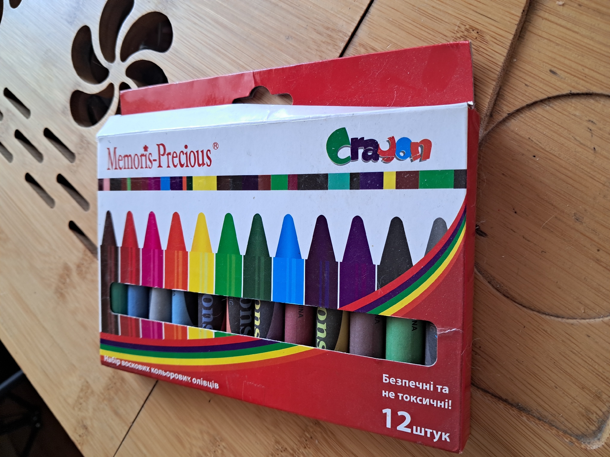A close-up, elevated shot shows a box of "Memoris-Precious" wax crayons resting on a light brown wooden surface with decorative cutouts. The crayon box, predominantly red and white, is positioned at an angle, revealing its front and top.
The top portion of the box is white, featuring the brand name "Memoris-Precious®" in red script on the left and the stylized word "Crayon" in green and blue on the right. Below this, a horizontal stripe displays small colored rectangles, mirroring the crayon colors inside. The main display area of the box features a row of twelve crayons with their pointed tips visible through a cut-out window. These crayons display a spectrum of colors including brown, red, pink, orange, yellow, green, dark green, blue, purple, dark purple, black, and light green. A rainbow stripe graphic runs along the bottom edge of the window.
Visible text on the box, below the crayons, is in Ukrainian. It reads "Набір воскових кольорових олівців" (Set of wax colored pencils/crayons) on the left, and "Безпечні та не токсичні!" (Safe and non-toxic!) followed by "12 штук" (12 pieces) on the right. Some individual crayons also show partial text like "ons" and "INA" (likely part of "Crayons" and "CHINA").
The wooden surface underneath and around the box has a distinct grain and carved, swirling patterns on the left, suggesting a decorative piece of furniture or paneling. The lighting indicates an indoor setting, likely during the daytime. The location of the image is Kyiv, Ukraine, which aligns with the Ukrainian text on the product packaging.