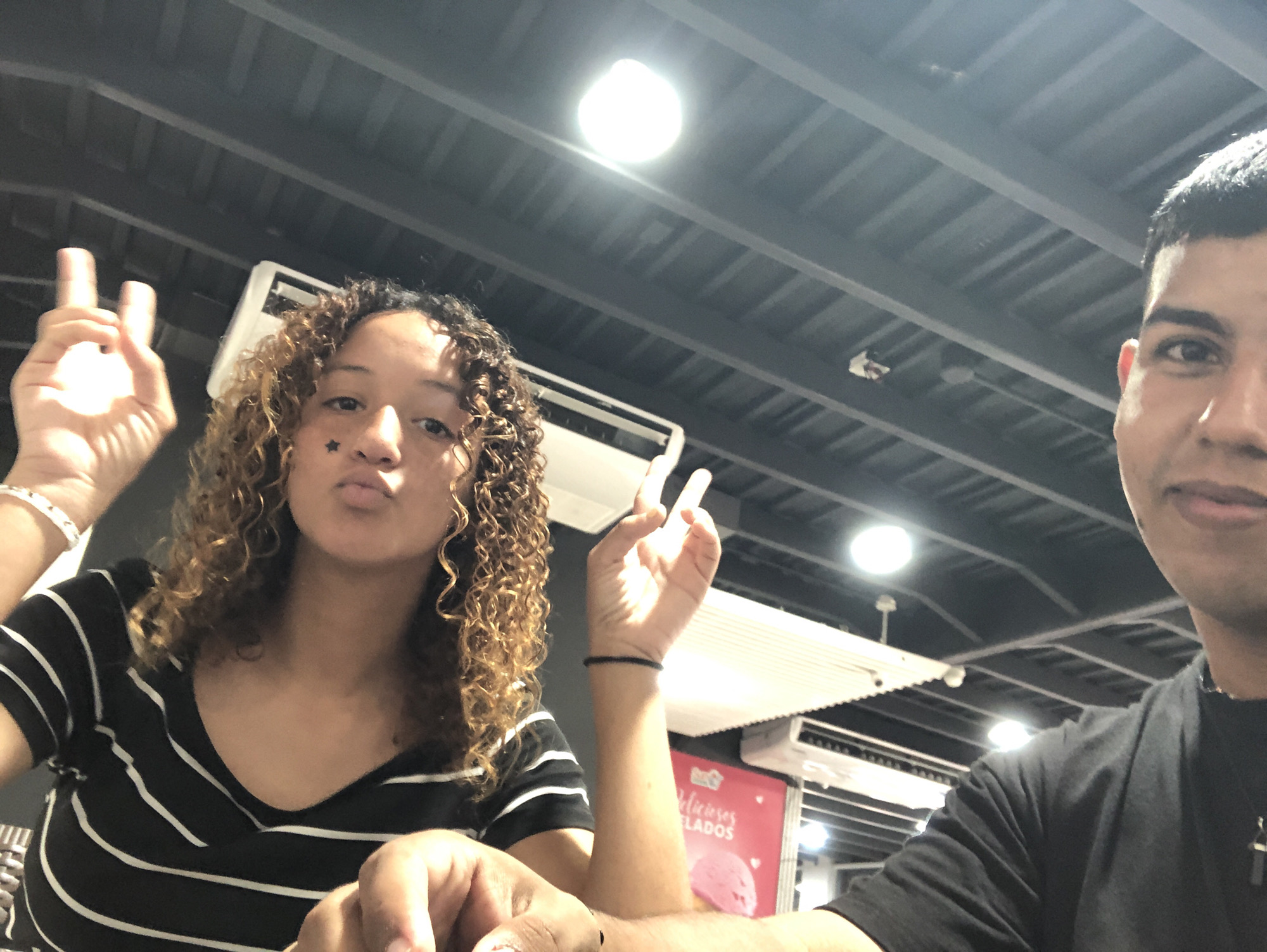 The image depicts two people in what appears to be an indoor setting, possibly a restaurant or commercial establishment. A woman with curly hair, wearing a black and white striped top, is making a peace sign with her left hand and puckering her lips. A star sticker is visible near her eye. A man, wearing a black shirt, is partially visible on the right side of the frame. He is looking at the camera. In the background, there is a ceiling with visible light fixtures, and air conditioning units. A pink sign with the words "Deliciosos Helados" is partially visible. The picture was taken in Juan Griego, Venezuela.