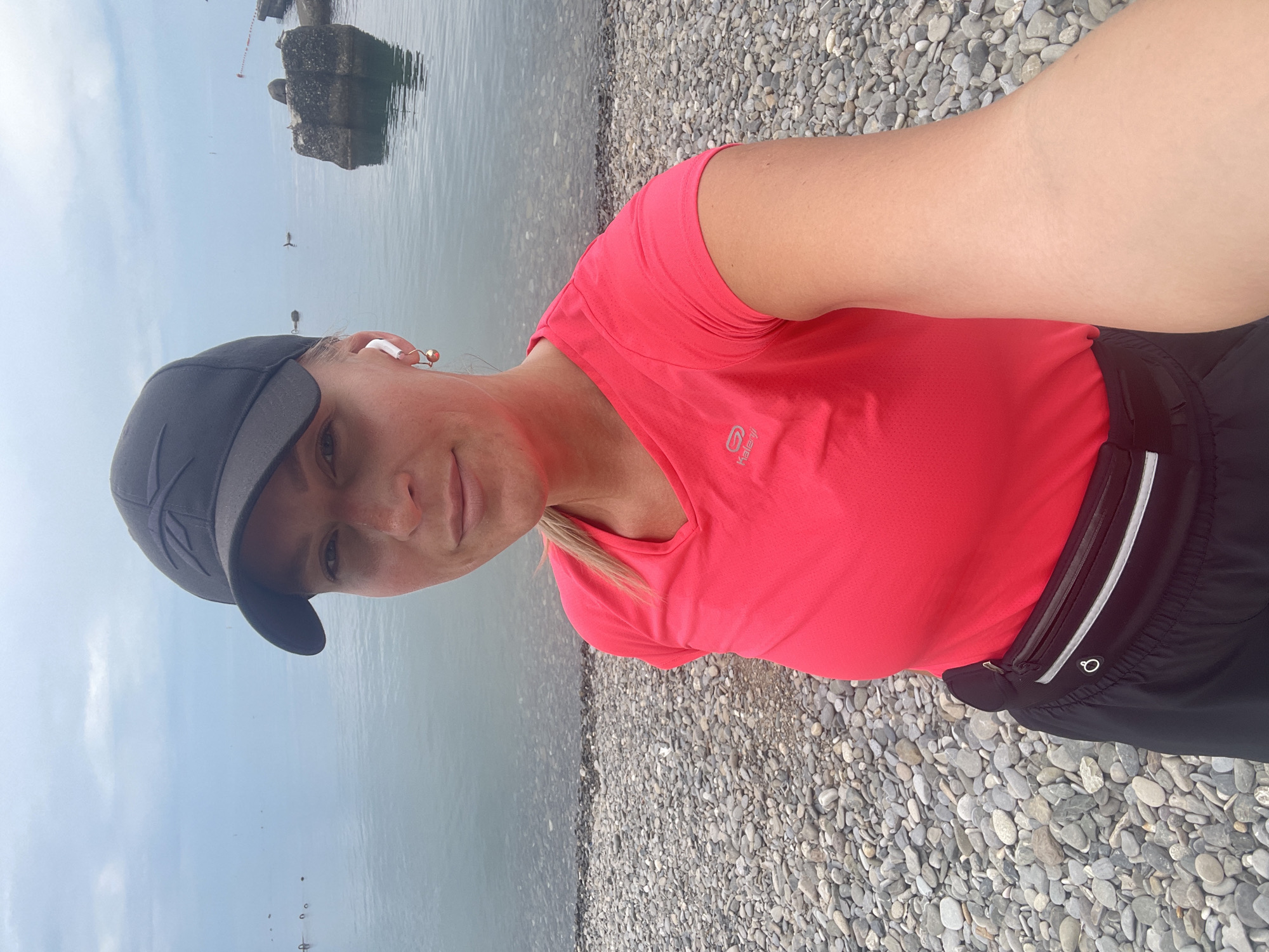 A woman stands on a pebble beach in Sochi, Russia, wearing a black baseball cap, a pink short-sleeved shirt, and black shorts with a waist pack. She has blonde hair and is wearing wireless earbuds. The background features a body of water, possibly the Black Sea, with some indistinct structures visible further out in the water. The sky is overcast.