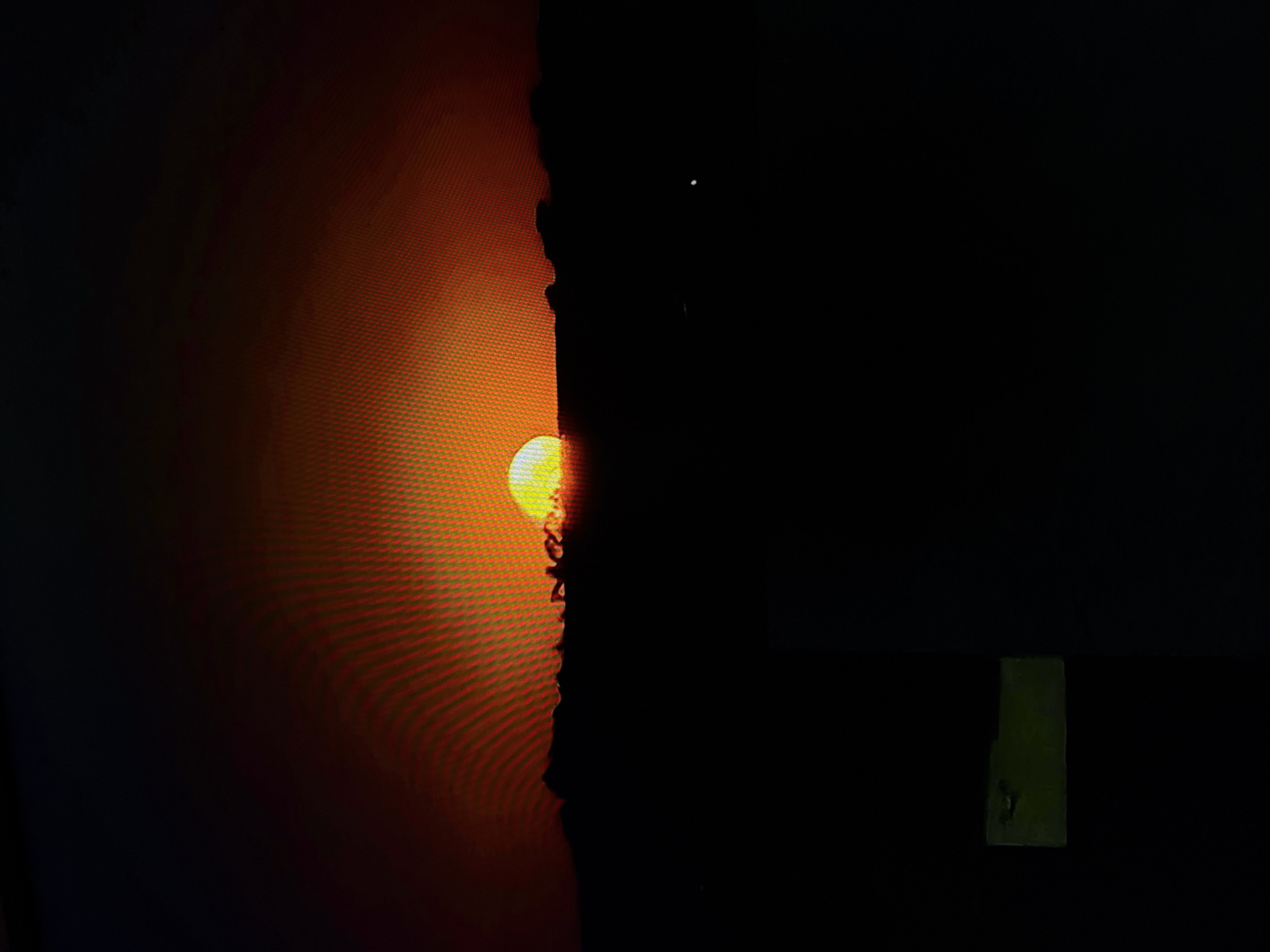 An image captures a sunset over a dark, silhouetted landscape. The sun, a bright orb of yellow and orange, is partially visible behind a dark horizon line, with rays of light fanning out across the upper portion of the frame. The sky transitions from a deep orange near the sun to a dark, almost black hue at the top. The lower half of the image is completely black, obscuring any details of the terrain or any potential objects. A small, faint white light is visible in the black expanse, likely a distant star or object. In the bottom left corner, a blurry, rectangular green object is partially visible against the black background.