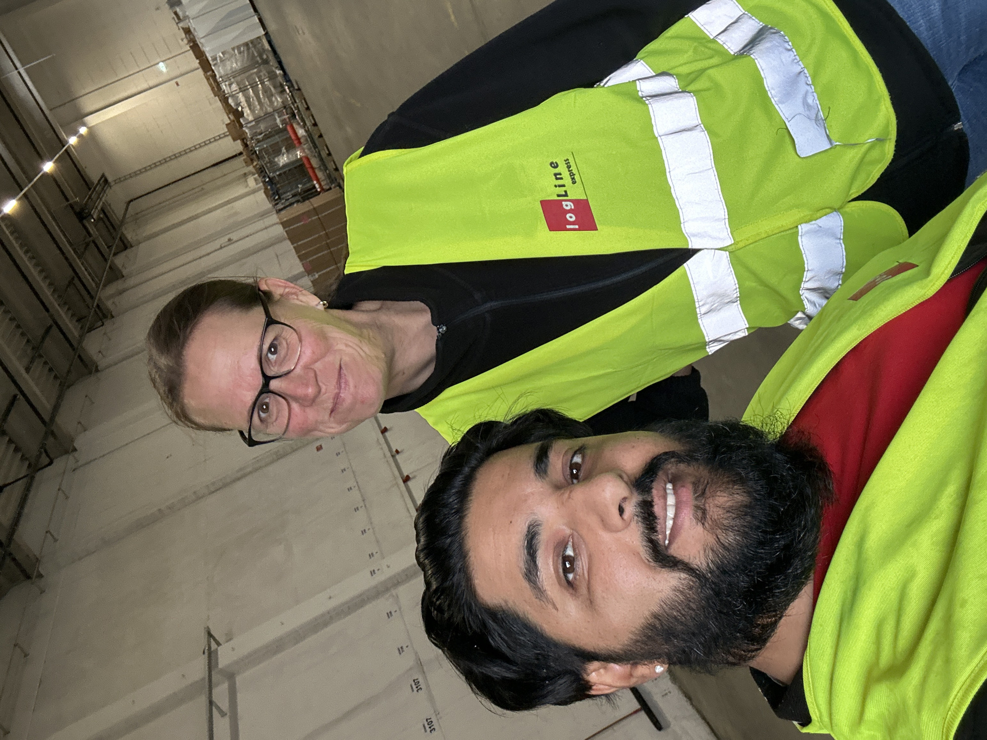 The image shows two individuals inside a warehouse environment. The person in the foreground is a man with dark hair and a beard, wearing a bright yellow-green safety vest over a red shirt. The person in the background is a woman with blonde hair and glasses, also wearing a yellow-green safety vest. The vest has a red and white logo that reads "logLine express". The warehouse appears to be large and industrial, with concrete walls and a high ceiling with lighting fixtures. Pallets of goods wrapped in plastic are visible in the background. The image was taken in Schweitenkirchen, Germany.
