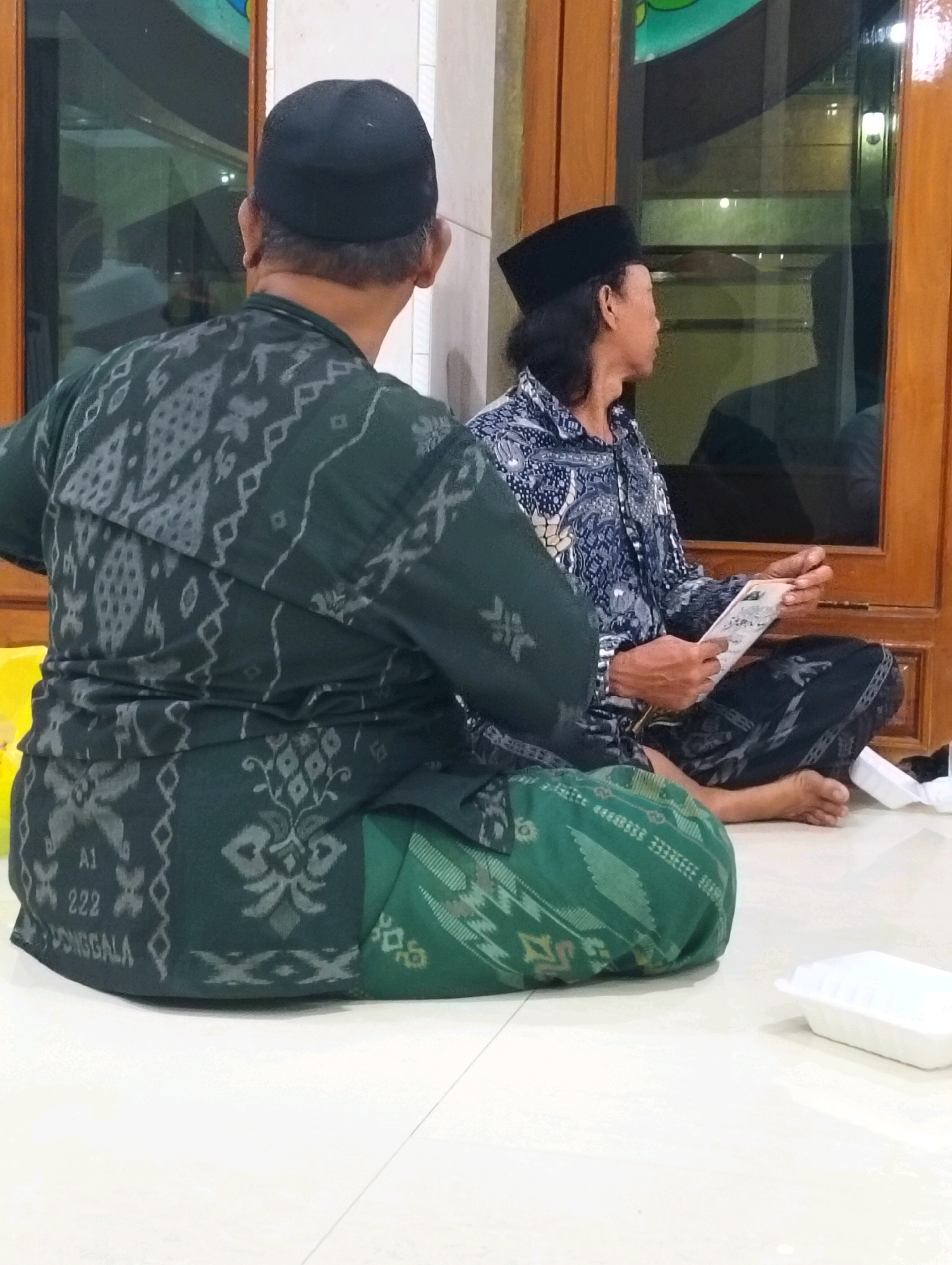 In the city of Dagan, Indonesia, two men are seated on the floor indoors, likely in a religious or community setting, judging by the traditional attire and the presence of what appears to be a prayer mat or similar textile. The man in the foreground, facing away from the camera, wears a dark green long-sleeved shirt with intricate grey patterns and a black cap. He is seated on a dark green and patterned textile. To his right, the second man, facing the side, wears a patterned blue and white shirt and a black cap. He is holding a folded paper document and is seated on a dark patterned textile. Both men are wearing sarongs, the traditional Indonesian garment. The environment includes a wall with wooden accents and large windows reflecting light and the interior. A white disposable food container is visible to the right of the seated men. The floor is tiled in a light color. The lighting suggests an indoor environment, possibly with artificial light sources.