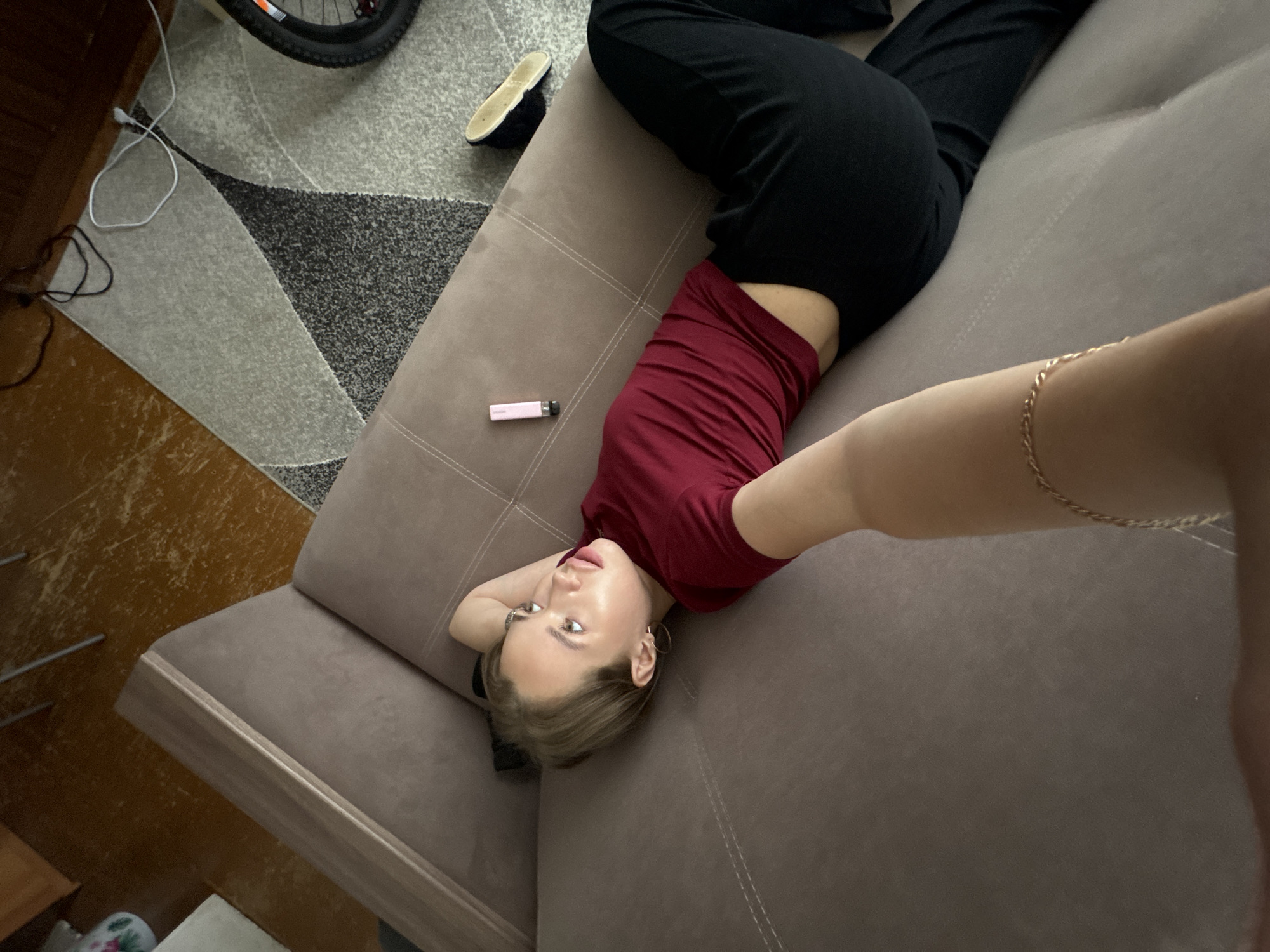 A young woman with blonde hair is lying on her back on a brown sofa. She is wearing a maroon t-shirt and black pants. Her head is propped up on her hands, and she is looking towards the ceiling. A pink vape pen and a single black slipper are on the sofa next to her. The room appears to be a living room, with a patterned rug on the floor and a wooden floor visible in the background. The image was taken in Chaikovsky, Russia.