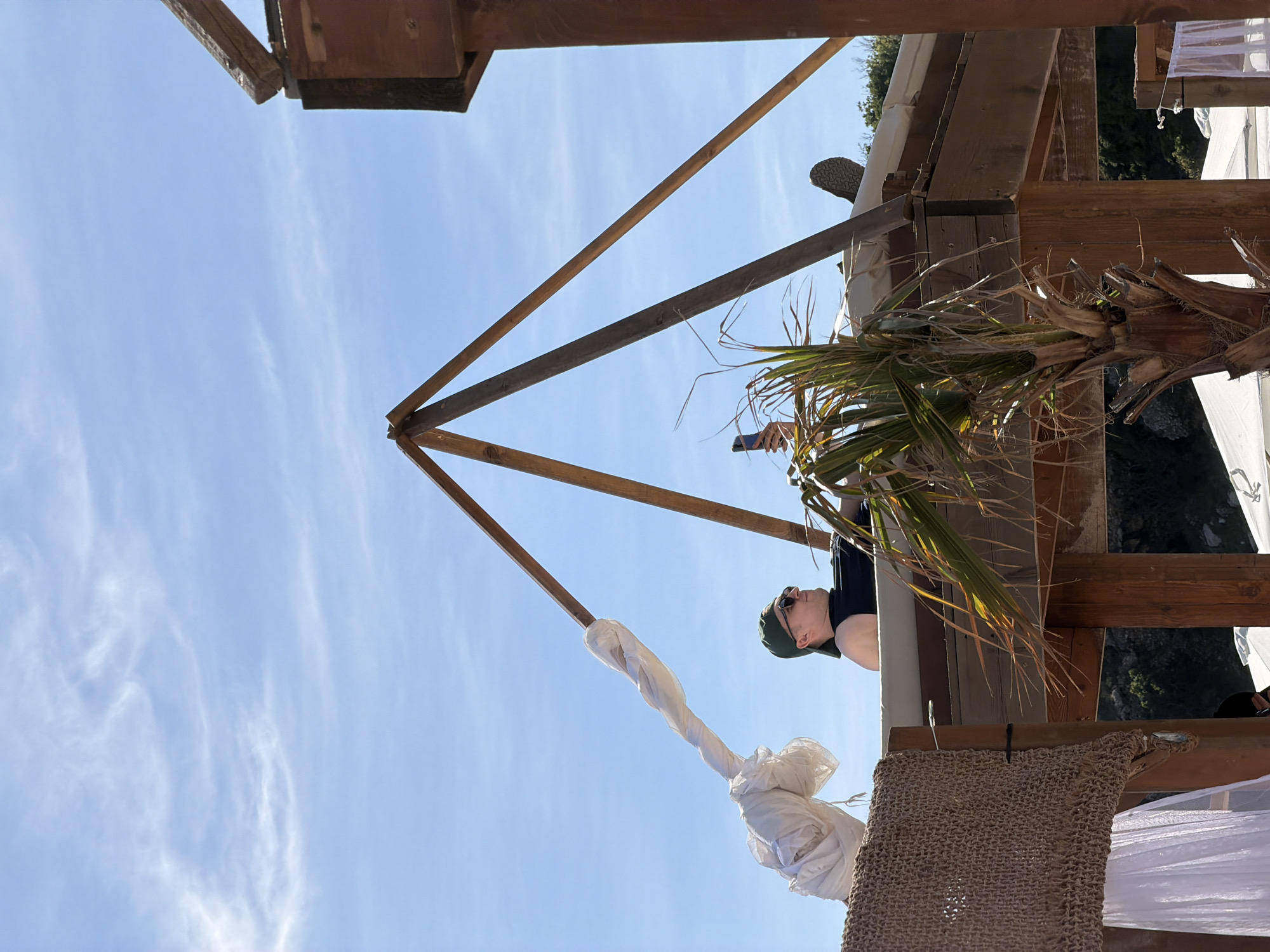 A low-angle shot reveals a person in sunglasses and a green cap, holding a smartphone, partially obscured by palm fronds. They stand on a wooden structure, presumably a cabana or beach bar, set against a backdrop of a clear blue sky with some wispy clouds. The structure has triangular wooden supports and decorative elements of white knotted fabric and woven netting. The location, inferred from the combination of architectural style and vegetation, could be Budva, Montenegro.