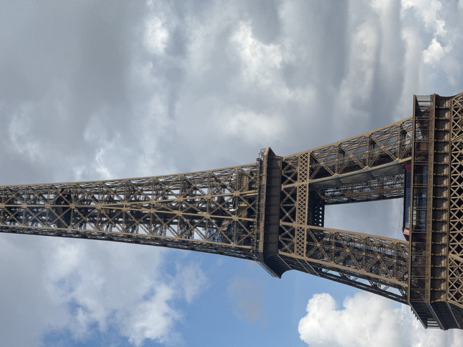 The image presents a detailed, vertical view of the lower and middle sections of the Eiffel Tower, located in Paris, France, against a mixed sky backdrop. The tower's intricate, dark bronze wrought-iron lattice structure dominates the frame, extending upwards from its wide base. The first major observation platform is clearly visible, featuring enclosed spaces with glass windows and a prominent reddish architectural element. Above this, the tower narrows, leading to the second public platform where numerous small human figures are discernible along the railing, indicating their presence as visitors. The sky on the left displays clear blue patches interspersed with white cumulus clouds, while the right side is characterized by an expanse of dense, grey overcast clouds. No other specific objects, actions, or direct interactions are clearly identifiable beyond the human presence on the platform.