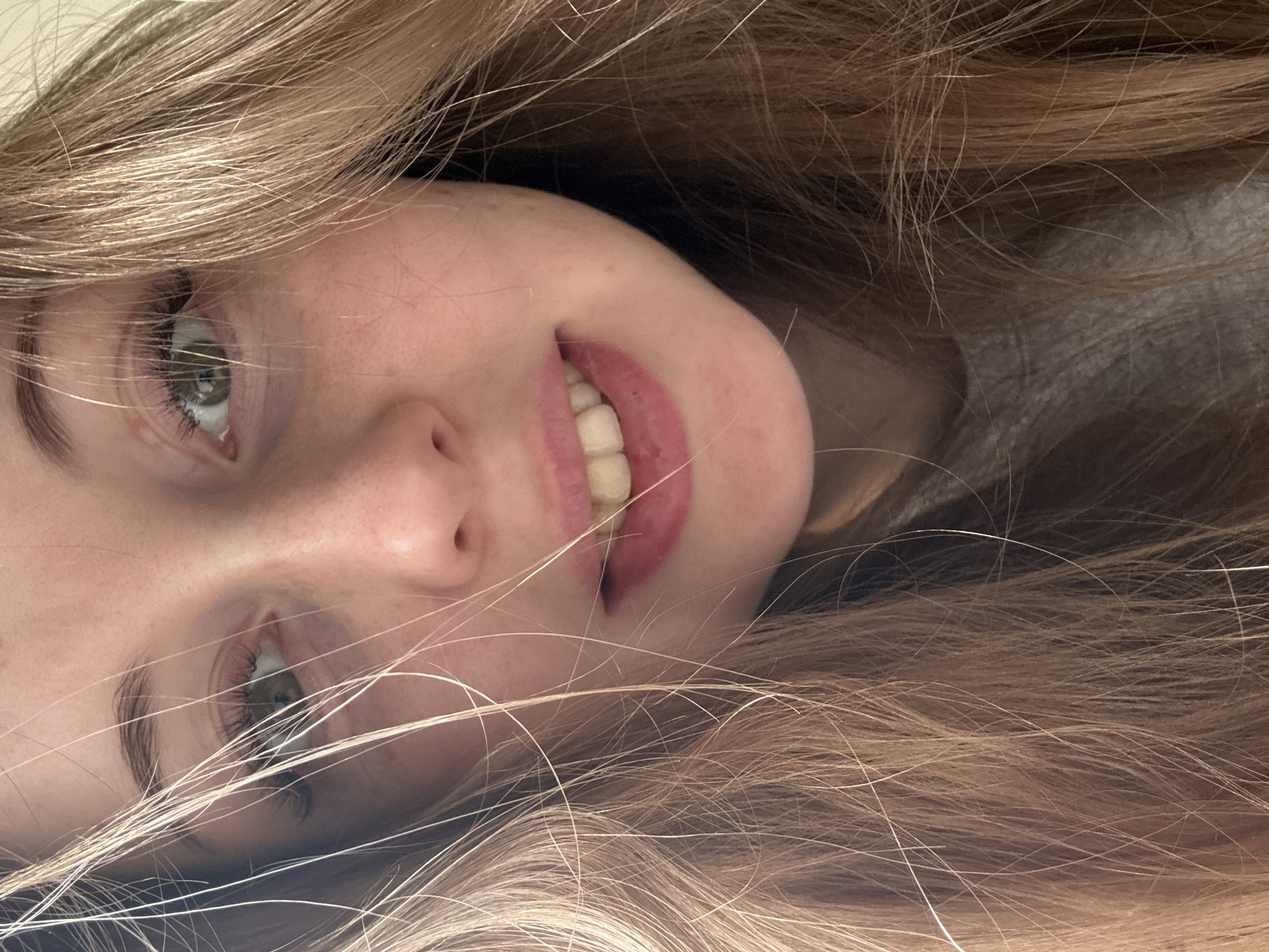 A close-up, rotated image depicts a young woman's face surrounded by strands of long, blonde hair. Her skin appears fair, with visible freckles. She is looking at the camera with grey-green eyes. Her lips are parted in a slight smile, revealing her teeth. The background is out of focus. The city location is unknown.