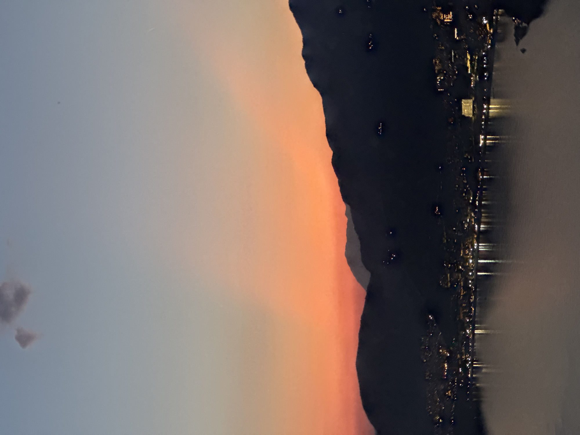 The image captures a view in Kamenovo, Montenegro, likely at dusk or dawn. A dark silhouette of a mountain occupies the central right area, with lights dotted along its base suggesting a settlement or road. These lights are reflected in the body of water to the far right, indicating a waterfront location. The sky in the left portion transitions from a light blue-grey to orange and red near the mountain ridge, suggesting a sunset or sunrise. A small cloud is visible in the left corner of the image.