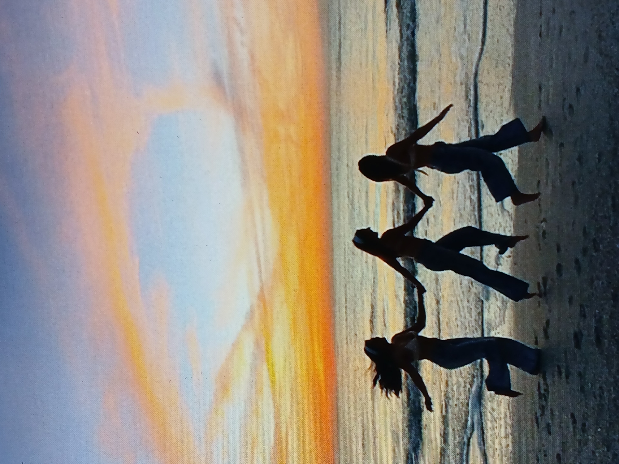 Three people are standing on a beach during a sunset. The location appears to be Juan Griego, Venezuela. The people are holding hands in a line. They are silhouetted against the bright sky. The beach is sandy and has waves breaking on it. The sky is a mix of orange, yellow, and blue.