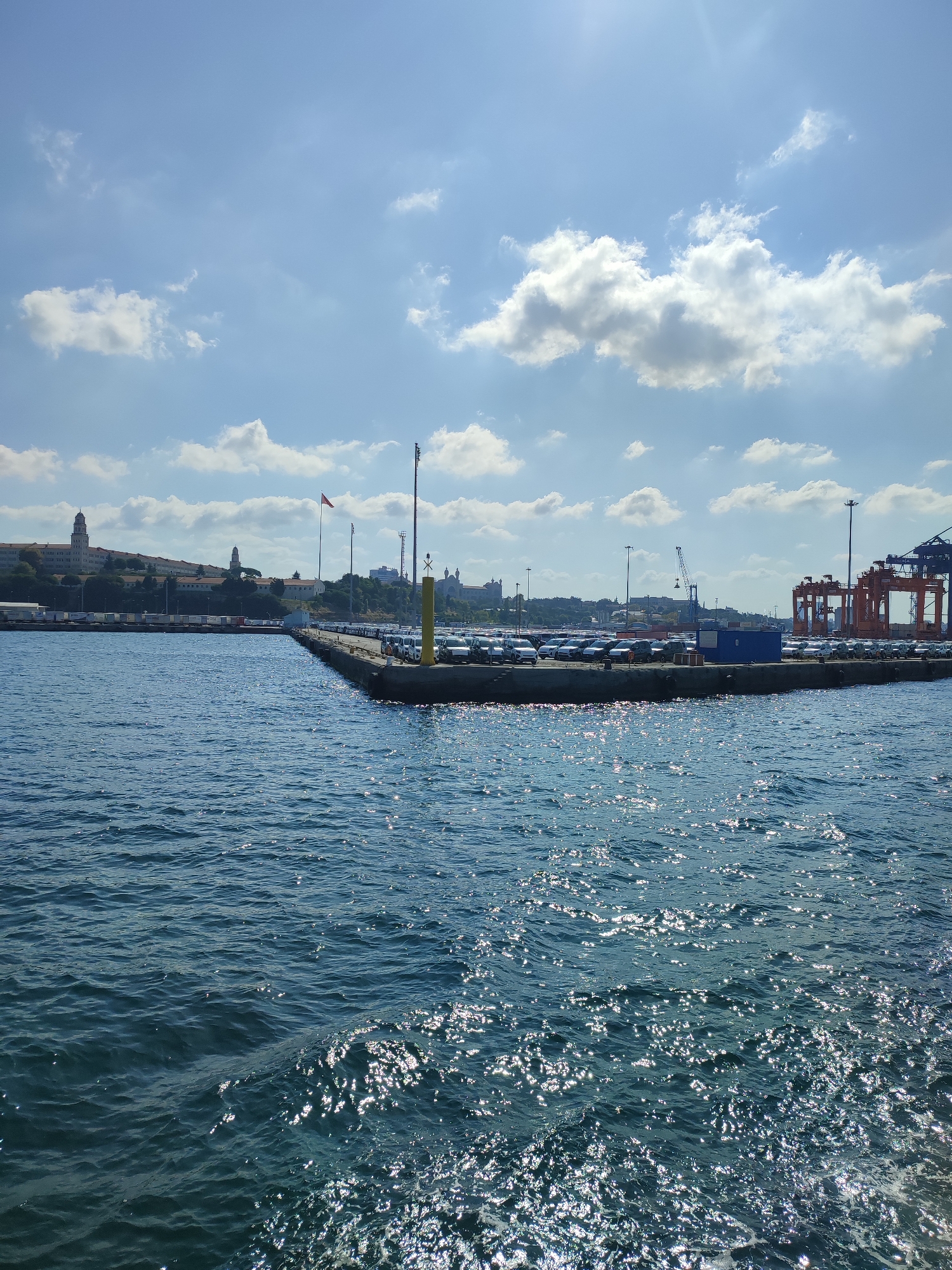 The image depicts a bright, sunny day at a port facility. The foreground is dominated by dark blue water, with sunlight reflecting off the surface, creating a shimmering effect. In the middle ground, a pier juts out into the water. The pier is lined with parked cars, appearing to be new or awaiting transport. A large blue shipping container sits on the pier. To the right of the pier, large orange shipping cranes are visible, indicative of a busy cargo terminal. In the background, a line of buildings, possibly administrative or residential, is situated on a gentle slope. One prominent building has a tall, cylindrical tower. A red flag flies from a flagpole on the pier. The sky is a vibrant blue with scattered white clouds. The scene suggests a functional port area, likely part of Üsküdar, Turkey, given its location on the Bosphorus Strait, a major shipping route. There are no people clearly visible in the image.