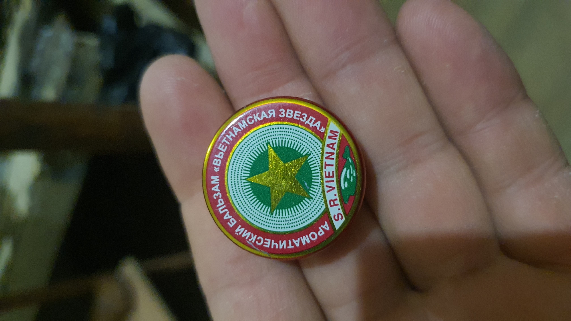 A person's hand is holding a small, round, metallic tin with a red and gold label. The label features a green circular design with a gold star in the center, surrounded by radiating lines. Text in a circular pattern reads "АРОМАТИЧЕСКИЙ БАЛЬЗАМ «ВЬЕТНАМСКАЯ ЗВЕЗДА»" and "S.R.VIETNAM". Below the circular design, there is a green band with a white, stylized depiction of a fish. The background is blurred and appears to be an indoor environment, possibly a room, with muted lighting. The image does not provide any information about the location in the city of Karankul, Uzbekistan.