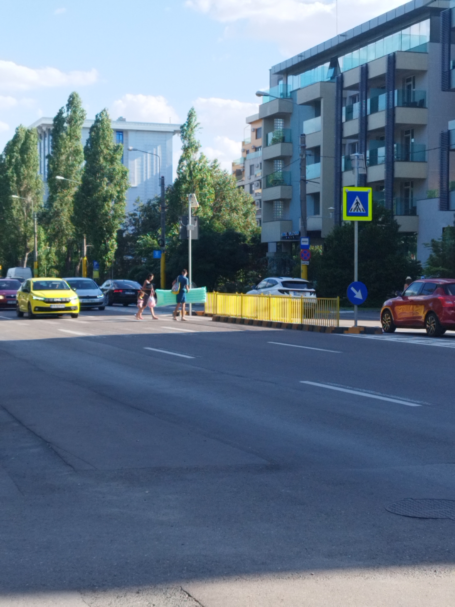 The image shows a street scene in Constanța, Romania, with a pedestrian crossing in the foreground. A yellow car is stopped at the crossing, and two pedestrians, a woman and a child, are walking across the street. A man is walking behind them. In the background, there are several buildings, trees, and traffic lights. The sky is blue with some clouds. The overall impression is of a typical urban environment on a sunny day.