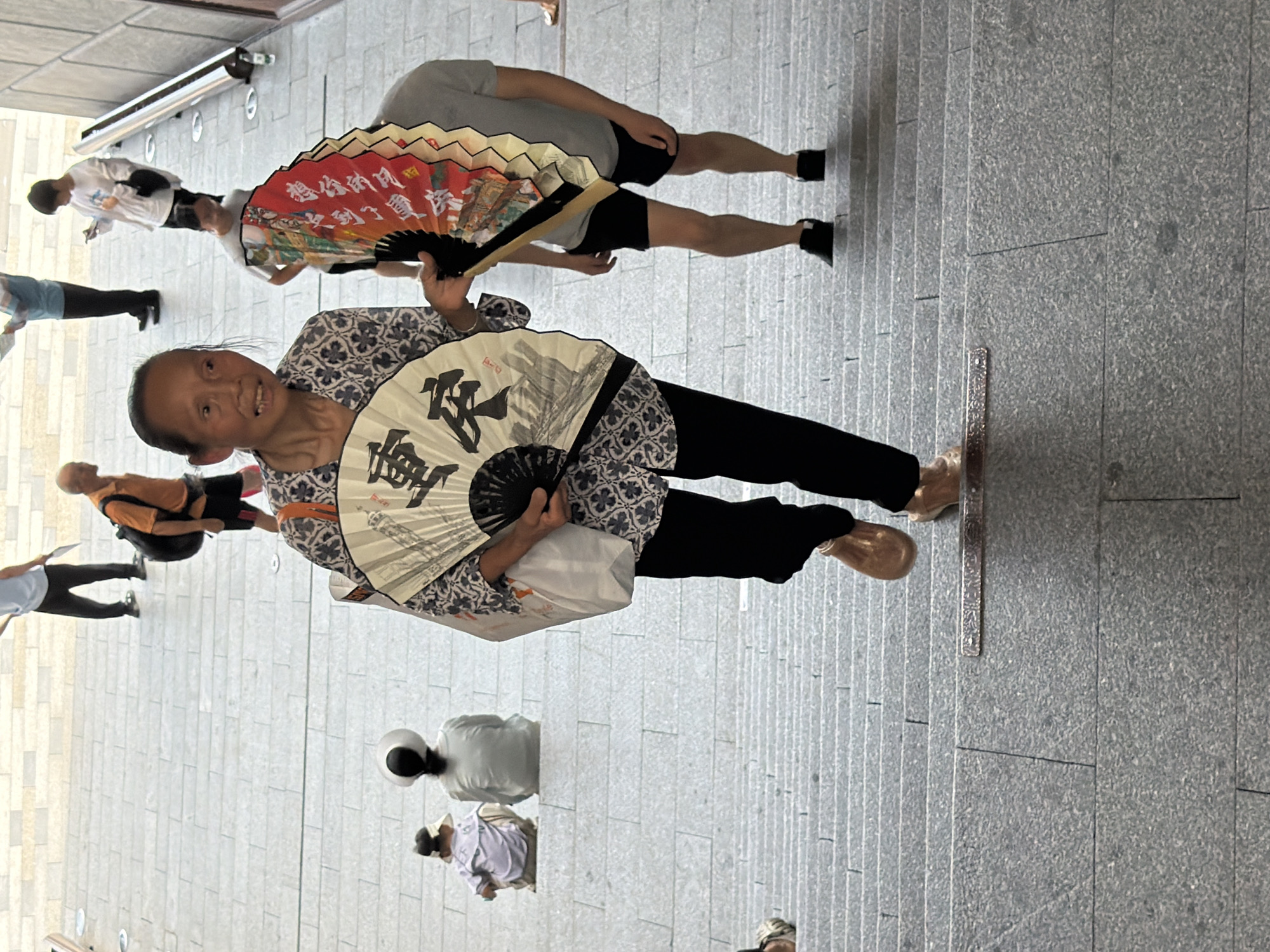 A street scene in the Yuzhong District of China features a woman holding two ornate fans, one white with black ink calligraphy and the other red with colorful imagery and text, as she walks up a wide stone staircase. The woman is in the foreground, facing slightly to her right, with her mouth open as if speaking. She wears a patterned blue and white blouse and black pants, with bronze-colored shoes. Behind her, other individuals are visible ascending and descending the stairs, including a person holding a red fan, another person in a grey t-shirt and black shorts, and two people seated on the stairs in the background. The environment consists of a grey, textured stone staircase with dark grout lines, suggesting an outdoor public space.