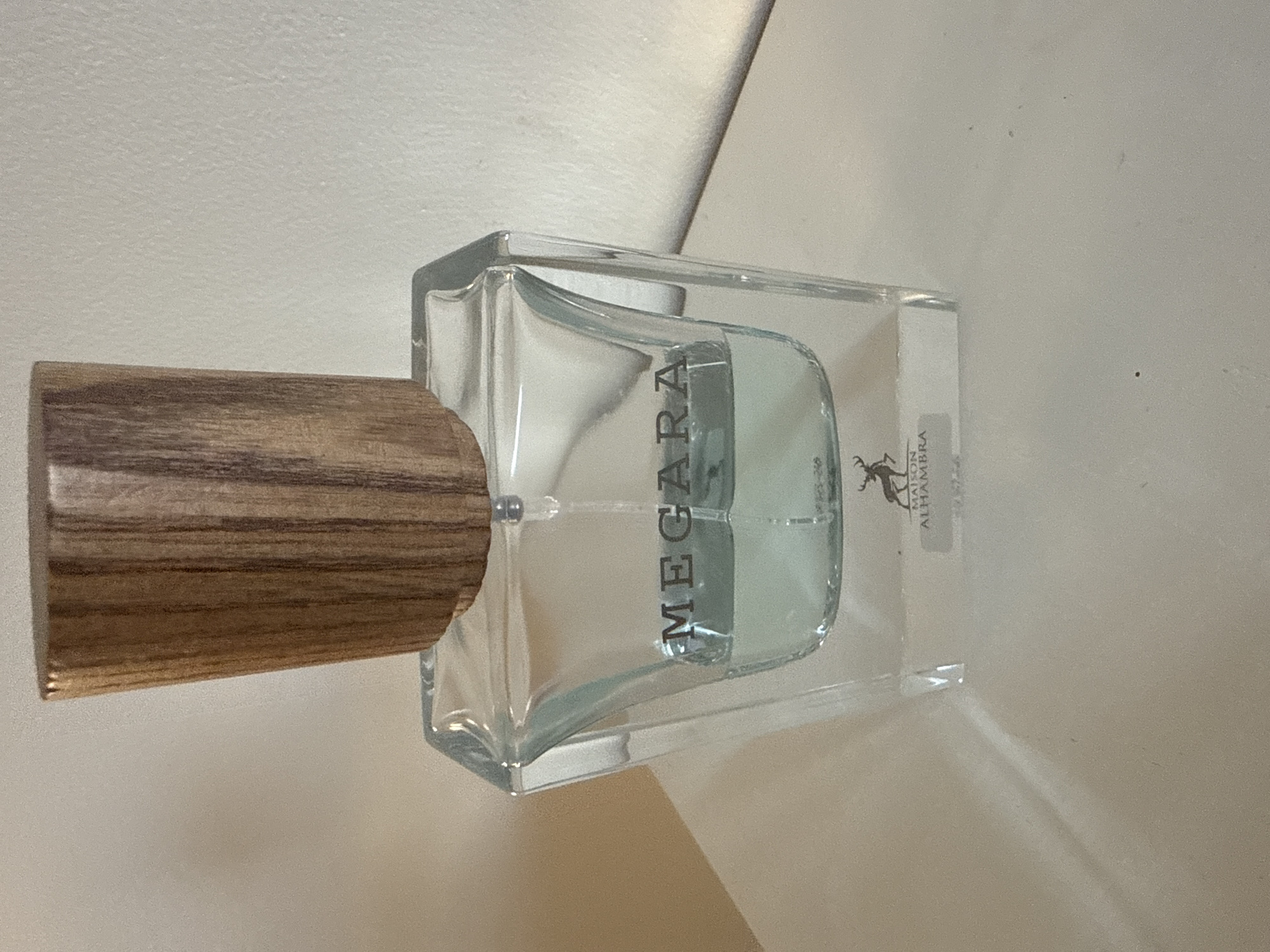 A clear glass perfume bottle with a wooden cap is the main subject of the image. The bottle contains a pale blue liquid and has the word "MEGARA" etched in dark lettering on its front. Below this, a rectangular label with a silhouette of a deer and the text "MAISON ALHAMBRA" is visible. The bottle is positioned on a flat, white surface, possibly a shelf or table, with a plain, light-colored wall as the background. The lighting in the image is soft, casting subtle shadows. The location in the city of Colchester, United Kingdom, is not determinable from the provided image.