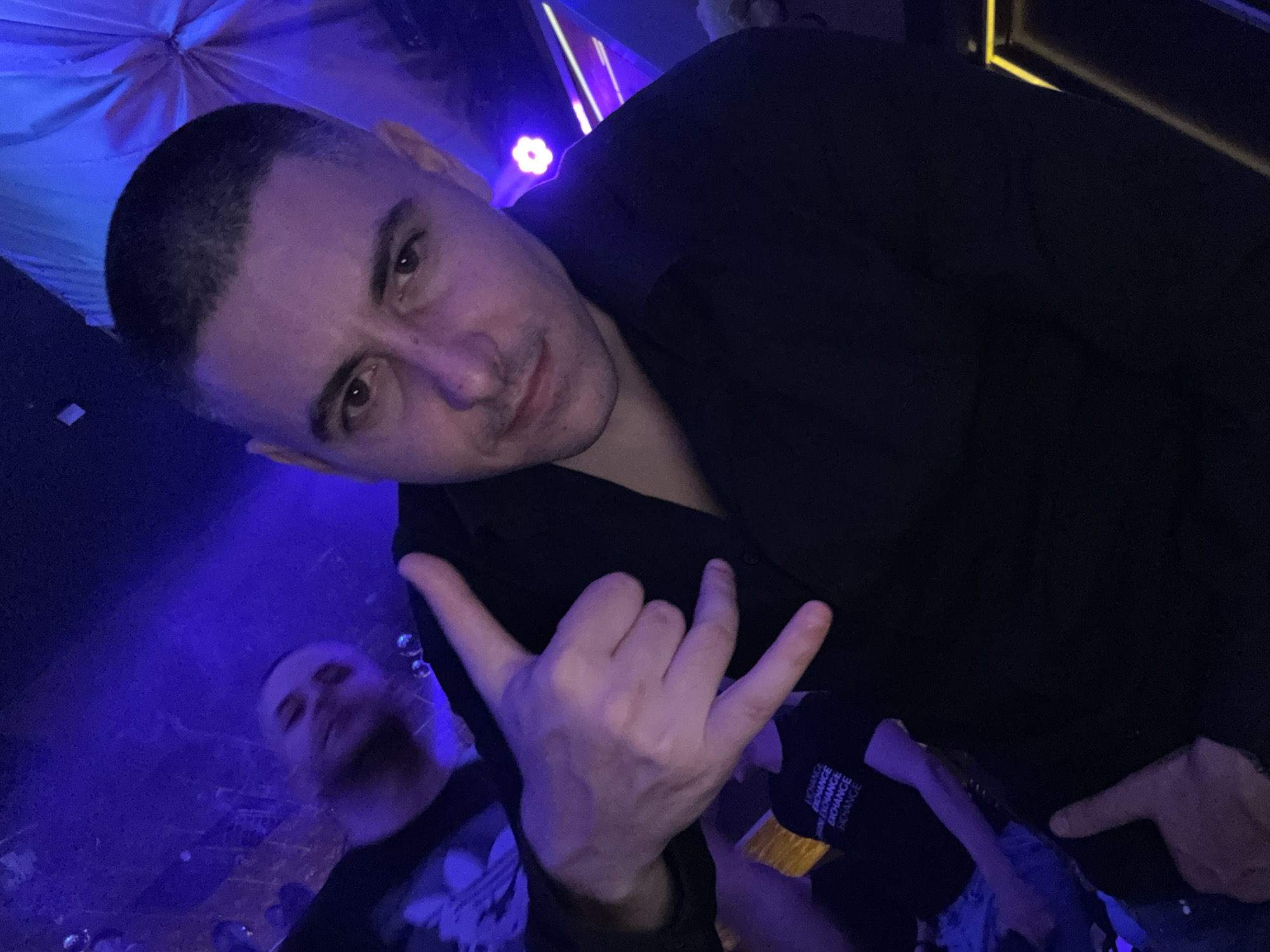 A man with a shaved head is in a club, making a "rock on" hand gesture. He is wearing a black shirt. Another man is partially visible in the lower left corner of the image. The club environment is illuminated with blue and yellow lighting. The location is in Pleven, Bulgaria.