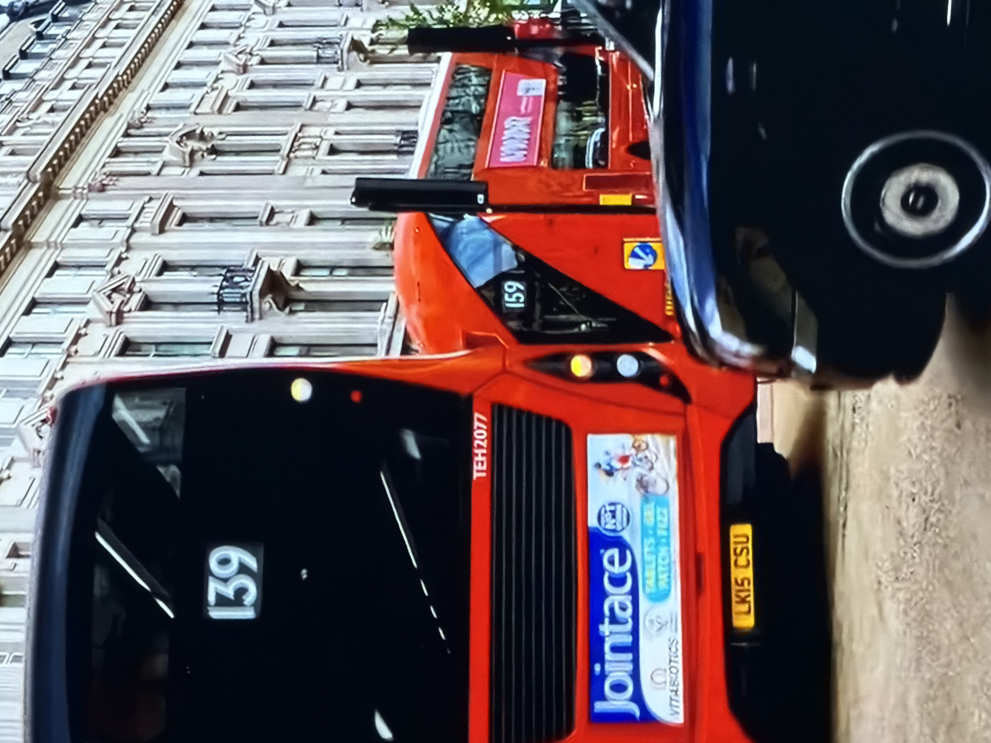 The scene is set in what is identified as Maidstone, United Kingdom, capturing an urban street with multiple vehicles and ornate architecture under daylight conditions.
In the foreground, the rear of a prominent red double-decker bus is visible. Its destination display shows the route number "139" in white digits on a black background. Below the rear window, the fleet number "TEH2077" is printed in white. A large advertisement for "Jointace" by Vitabiotics, featuring the text "TABLETS - GEL - PATCH - FIZZ" and depicting a person on a bicycle, is displayed across the lower rear section. The bus's license plate, located centrally at the bottom, reads "LK15 CSU" on a yellow background. The bus features horizontal black grille lines above the advertisement and has circular rear lights on its right side, including an amber indicator and a white reverse light.
To the right of the main bus, partially obscured, is another red double-decker bus, seen from its front-right side. It displays the route number "159" on its front destination board. A red advertising banner with indistinct white text, possibly "NOW", is visible on its side. A black vertical pole stands between the two buses.
In the immediate bottom-right foreground, a dark-colored vehicle, possibly a black or dark blue car or taxi, is partially visible, showing its side profile and a distinct silver-rimmed wheel.
The background features a long line of multi-story, ornate buildings, likely constructed from light-colored stone or stucco. These buildings exhibit classical architectural details, including multiple rectangular windows, some with decorative lintels and sills, and several small balconies with black wrought-iron railings. Decorative cornices and friezes are visible between floors and along the roofline, contributing to a grand urban streetscape. The street surface is paved.