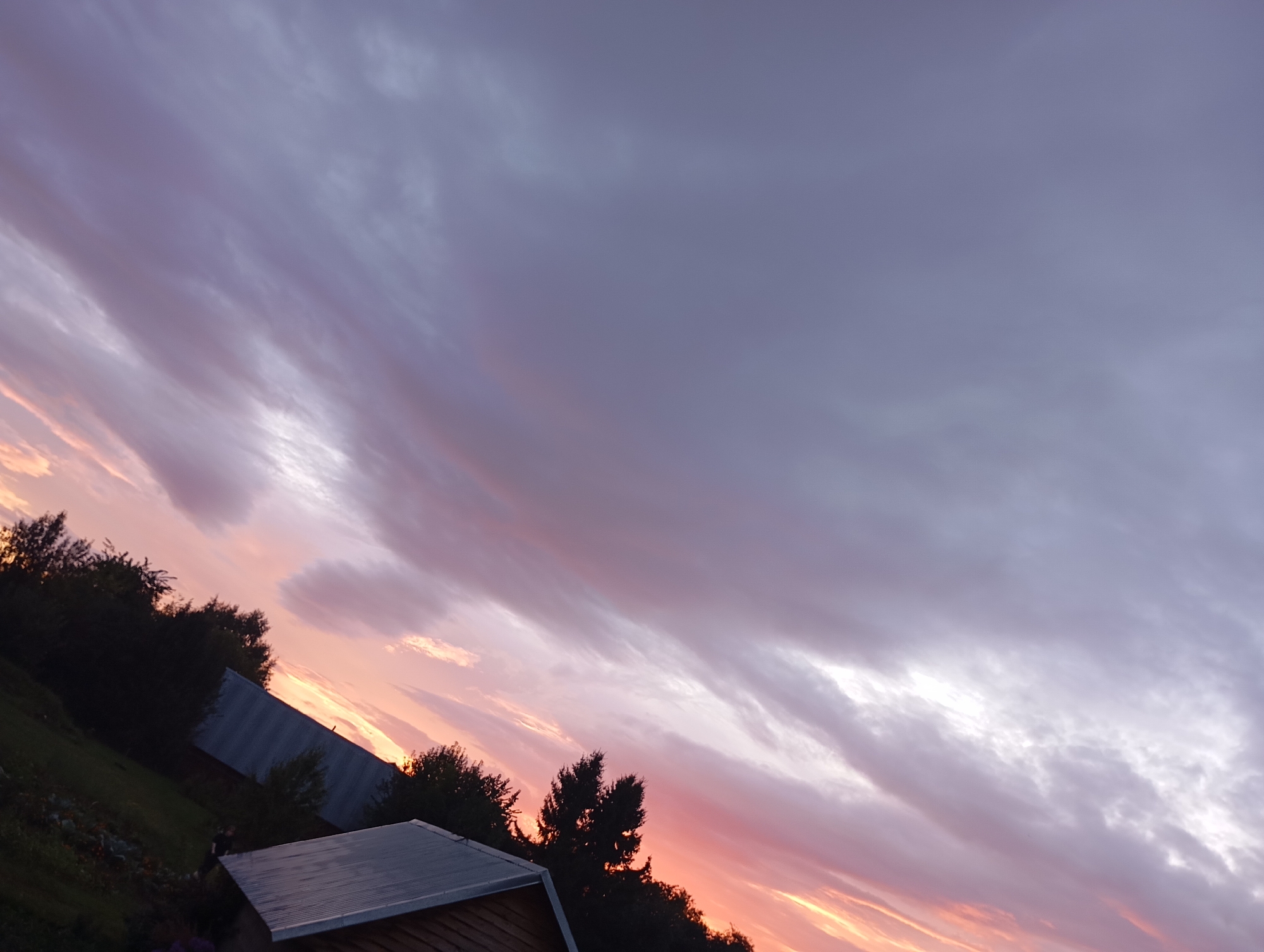 The image is a slightly tilted, wide-angle shot of a sunset over a rural area. The sky is filled with dramatic clouds displaying a gradient of colors from deep purple at the top to vibrant pinks and oranges near the horizon. Silhouetted trees and parts of buildings, likely rural structures such as sheds or small houses with metal roofs, are visible in the lower portion of the frame. The overall atmosphere suggests a serene, natural landscape at dusk. The location in Фоки, Russia, is not visually identifiable within the image itself.