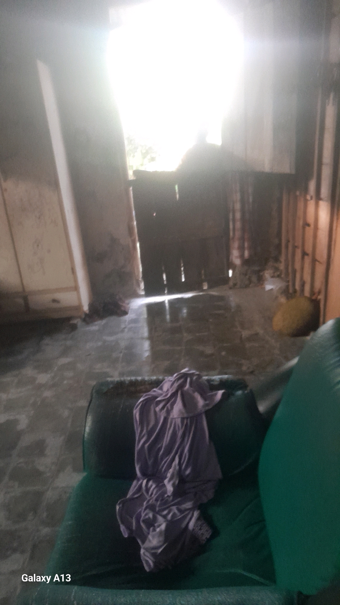 The image depicts an indoor scene, likely a room in a dwelling, with a distressed and somewhat neglected appearance. The lighting is very bright, originating from an open doorway or large window at the far end of the room, creating a strong silhouette and overexposure in that area.
In the foreground, a dark green couch or sofa is visible, with a piece of light purple fabric, possibly clothing, draped over its left side. The fabric is creased and loosely folded.
The floor appears to be tiled or a similar hard surface, with a sheen suggesting it might be wet or polished, reflecting the light from the doorway. The walls show signs of wear, with some areas appearing discolored or damaged. To the left of the doorway, a section of a wall or furniture piece is lighter in color.
The doorway itself is dark, with vertical elements suggesting wooden planks or a slatted structure. Beyond the doorway, foliage is visible, indicating an outdoor environment. To the right of the doorway, a cylindrical object with a yellow hue is present, possibly a large ball or cushion.
The overall impression is one of an interior space that is either undergoing renovation, has been abandoned, or is simply in a state of disarray. There are no people clearly discernible in the image, though the bright area at the doorway might contain a silhouette. The scene appears to be located in Ngelo, Indonesia, as indicated by the visible text "Galaxy A13" which is a brand of smartphone.