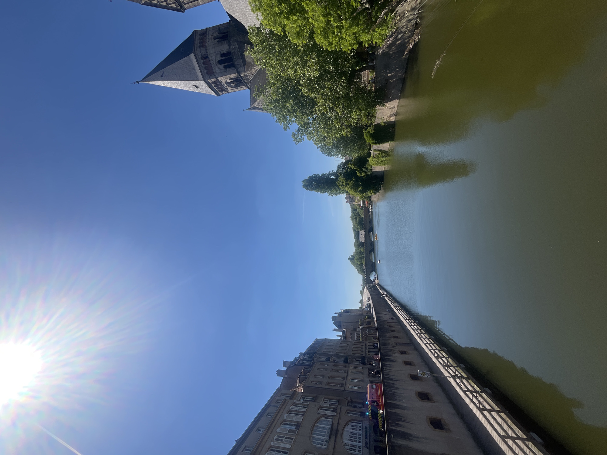 The image captures a sun-drenched scene in Metz, France, along the Moselle River. The Basilica of Saint-Pierre-aux-Nonnains, identifiable by its distinctive tower, is visible to the north. A long, stone embankment runs along the river's edge, lined with metal railings. Buildings, primarily residential, stand adjacent to the embankment, with a fire engine parked along the street. The river reflects the clear blue sky and the surrounding architecture. Trees line the riverbank. The sun is positioned to the west, casting bright rays across the scene.