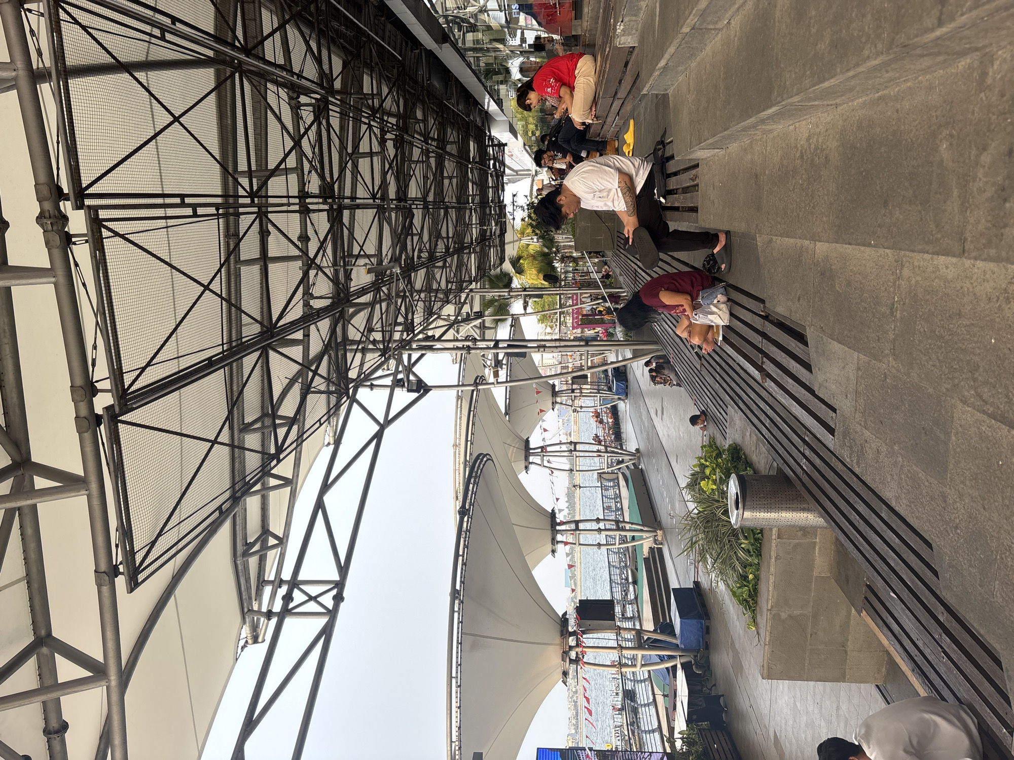 The image depicts an outdoor seating area with a large, modern canopy structure at a waterfront location in North Jakarta, Indonesia. Several individuals are seated on tiered wooden benches, many of whom are engaged with their mobile phones. In the foreground, a person wearing a white t-shirt and black pants is looking at their phone. To their left, a person in a maroon top is also looking down. Further back, a woman in a red top and light-colored pants is using her phone, and behind her, another person is seated. The background reveals a waterfront with boats and a cityscape in the distance. The architecture features a metal framework supporting large, white fabric canopies, and the seating area is constructed with stone and wood. A trash receptacle is visible near a planter with greenery. The lighting suggests it is daytime, with the sky being overcast.