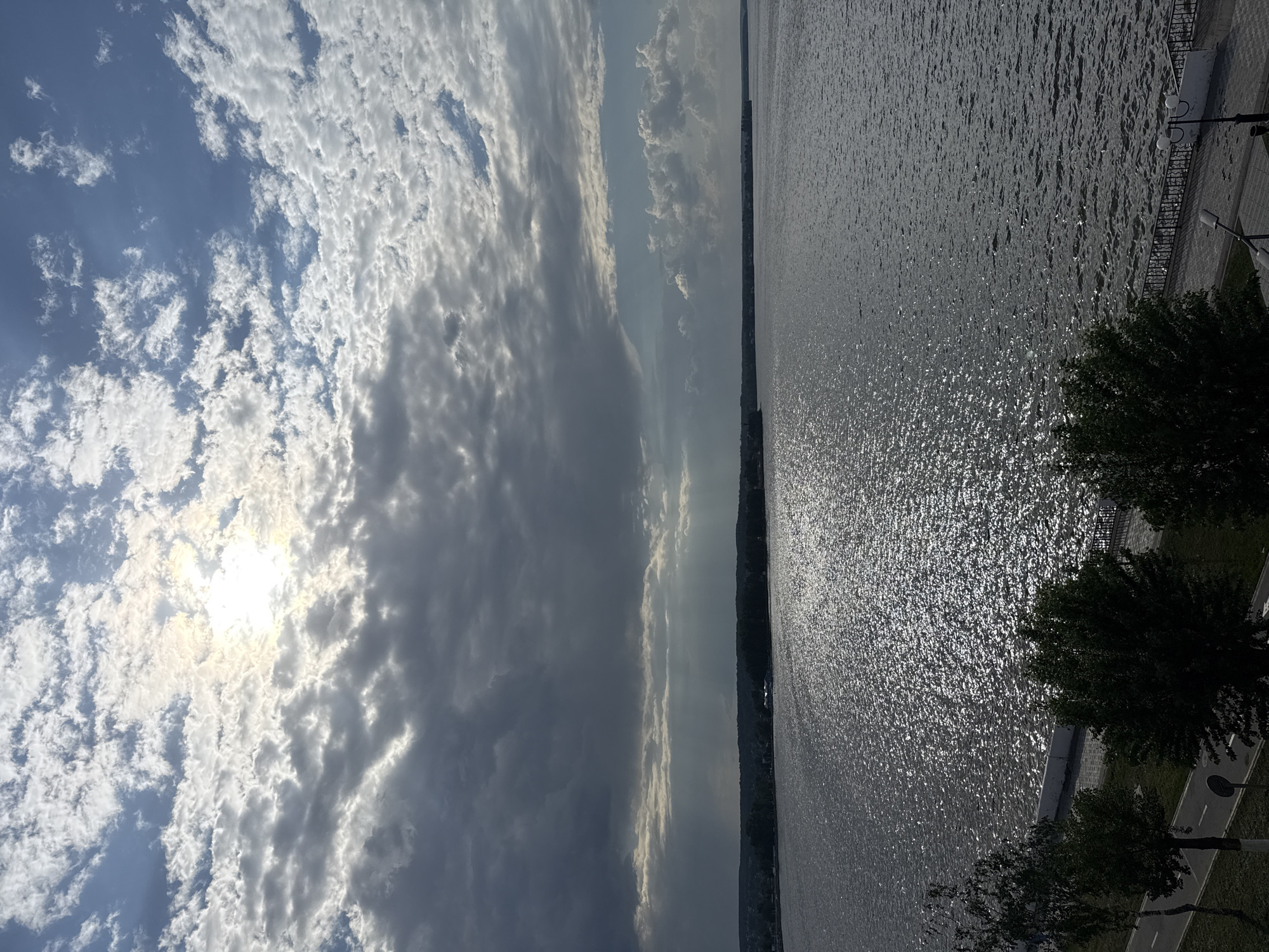 The image shows a body of water, likely a lake or wide river, under a sky filled with clouds. The sun is visible behind the clouds, creating a bright spot in the sky. The water surface is reflecting the sunlight, creating a shimmering effect.
On the right side of the image, there is a park or embankment with trees, sidewalks, and streetlights.
Based on the information provided and the visual elements in the image, it is likely that this scene is located in Votkinsk, Russia, and features the Votkinsk Reservoir.