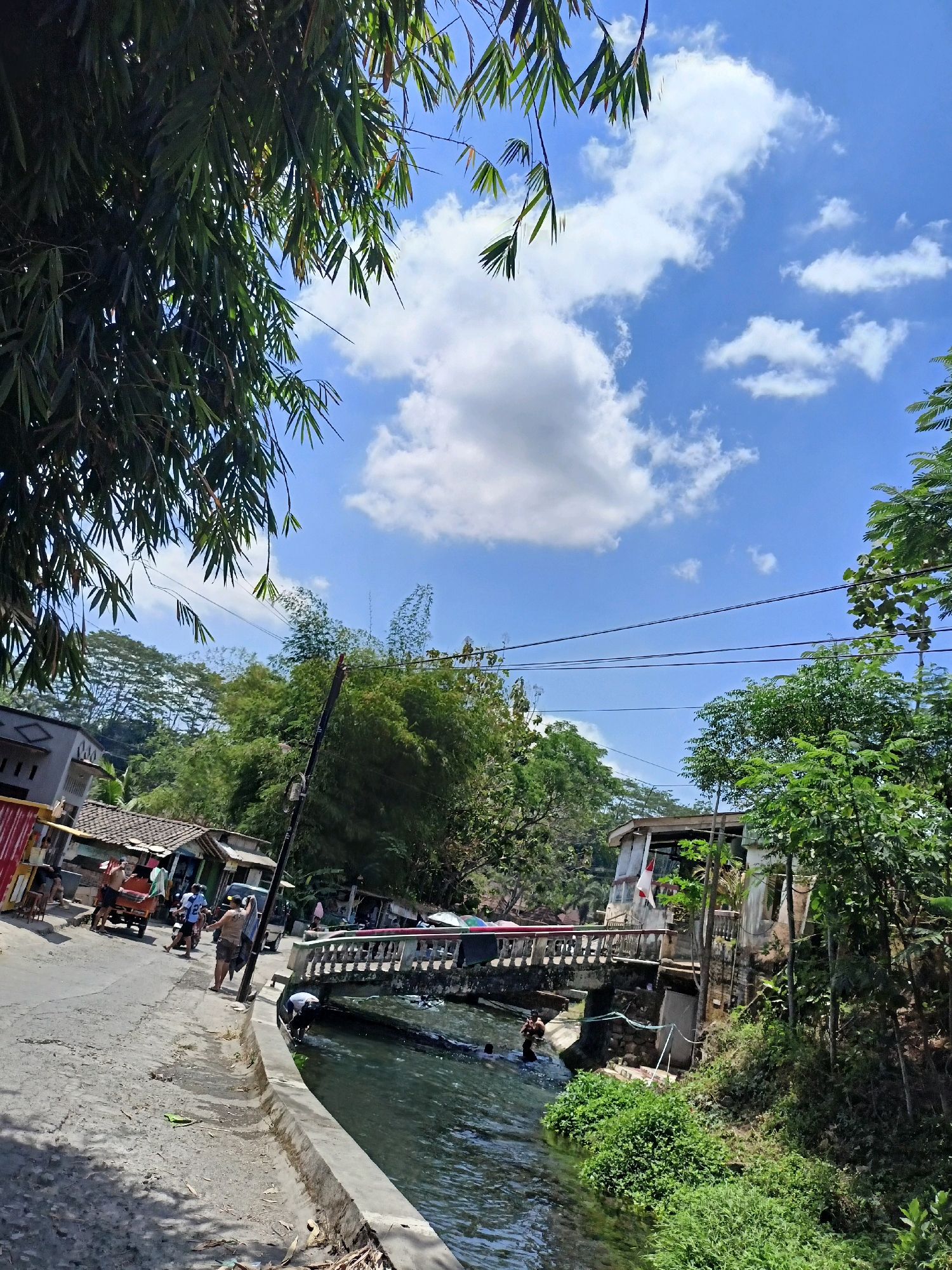 The image depicts a sunny day in Payaman, Indonesia, with lush green trees and a bright blue sky filled with fluffy white clouds. A paved road curves through the scene, with several buildings and structures lining its sides. On the left side of the road, a two-story building with a grey tiled roof is visible, along with smaller structures with tiled roofs. A cart pulled by a person is on the road, and a few individuals are walking.
A bridge spans a body of water, which appears to be a river or canal, running alongside the road. Some people are in the water, some swimming, and one person is bending down near the edge of the water. The bridge has a decorative railing, and a building with a light-colored facade and a flag is situated on the right side of the bridge. Dense vegetation, including bamboo, frames the upper portion of the image and the right side of the scene. Electrical wires are strung across the sky. The overall atmosphere suggests a lively, outdoor environment, possibly a village or a recreational area.