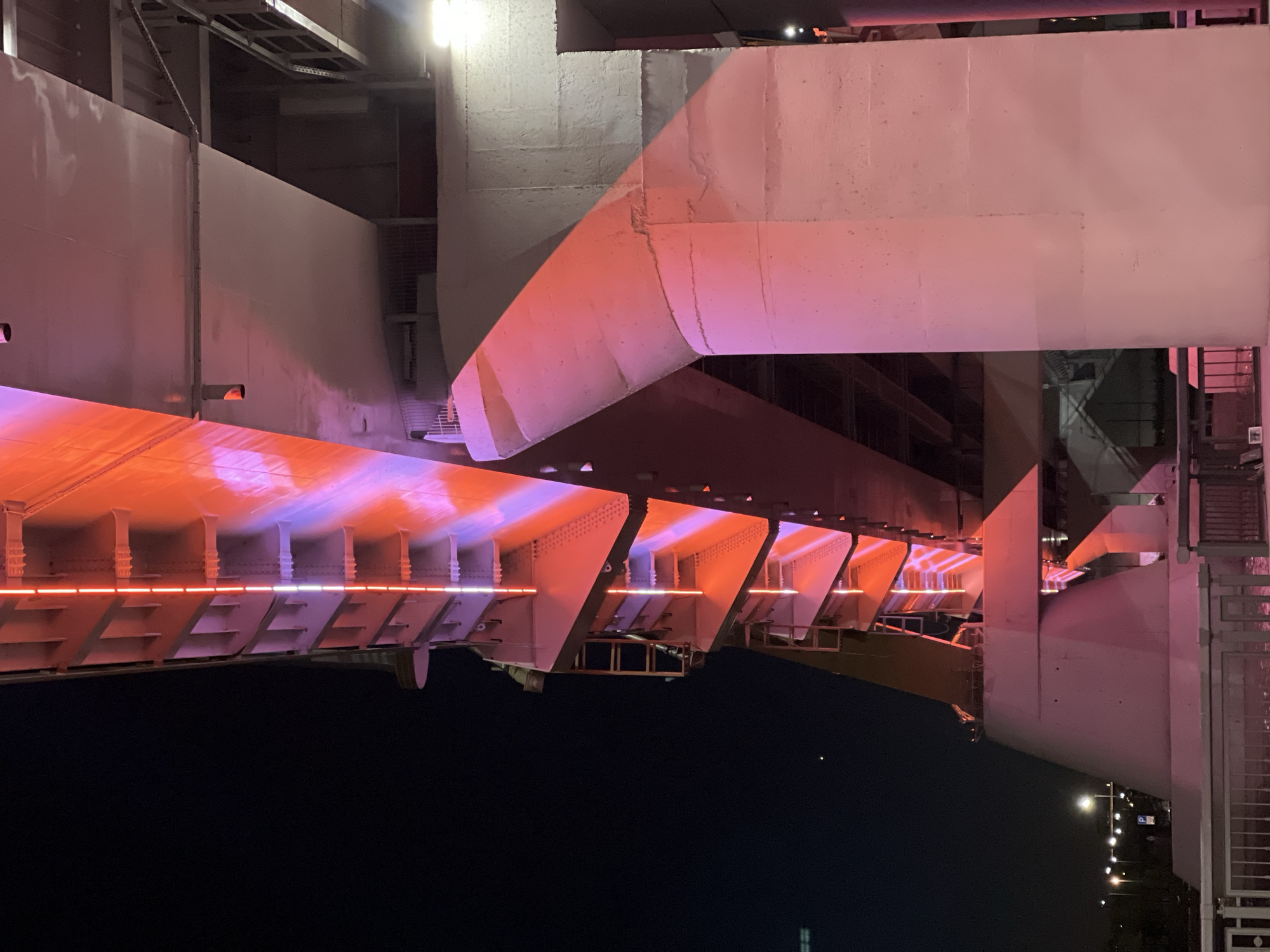 The image depicts the underside of the Milennium Bridge in Kazan, Russia, at night. The bridge's structure is illuminated with orange and purple lighting. The sky is dark. The supporting girders and structural elements of the bridge are visible. Streetlights are visible in the background.