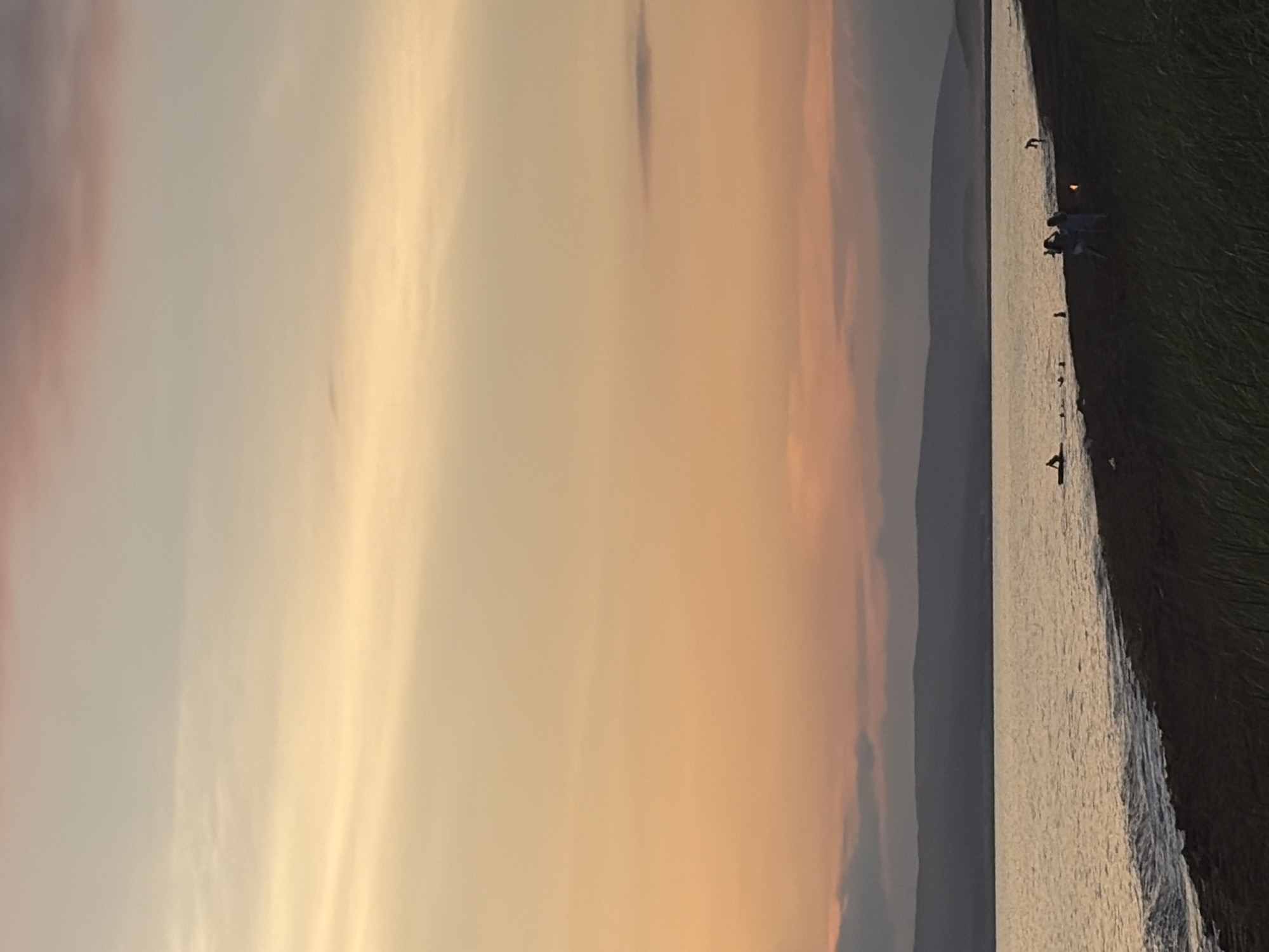 A sunset is captured over the body of water in Ozerskoe, Russia. The sky transitions from a soft peach and orange near the horizon to a pale yellow and gray higher up. In the distance, dark, undulating hills form the horizon line. The water is choppy with small waves lapping at a dark sand beach. Several people are present on the beach, some walking and others near the water's edge. One individual is visible on a board in the water. Green grass can be seen in the foreground, suggesting the edge of a park or natural area.