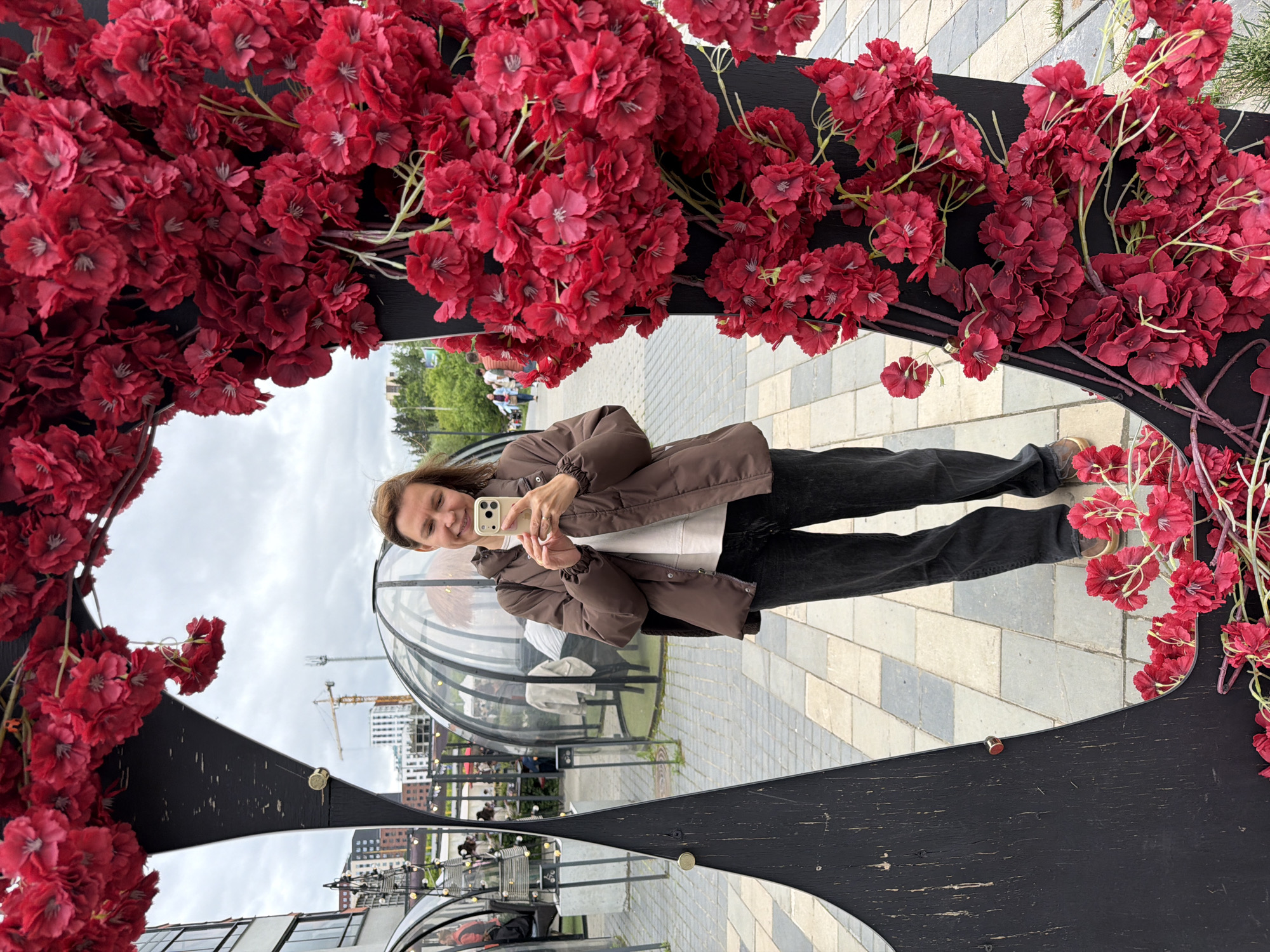 An outdoor scene in Izhevsk, Russia, depicts a middle-aged woman taking a selfie in a large, irregularly shaped, matte black mirror adorned with numerous clusters of artificial red flowers. The woman, with brown, shoulder-length hair and a smiling expression, is reflected from the waist up. She wears a brown puffer jacket over a white long-sleeved top, dark grey loose-fitting trousers, and brown shoes. She holds a light-colored smartphone with a triple-camera array in both hands, positioned to capture her reflection. The black mirror frame exhibits wear, with visible scratches and two metallic studs on its left edge.
The surrounding environment consists of a paved area with light and dark grey square and rectangular pavers. In the background, several transparent, dome-shaped outdoor dining pods are visible, some containing tables, chairs, and indistinct figures or objects. Beyond these, modern buildings with dark facades and glass elements are seen on the left. A yellow construction crane stands against an overcast, light grey sky further in the distance. Green foliage, including trees and bushes, is visible on the horizon, particularly to the right.