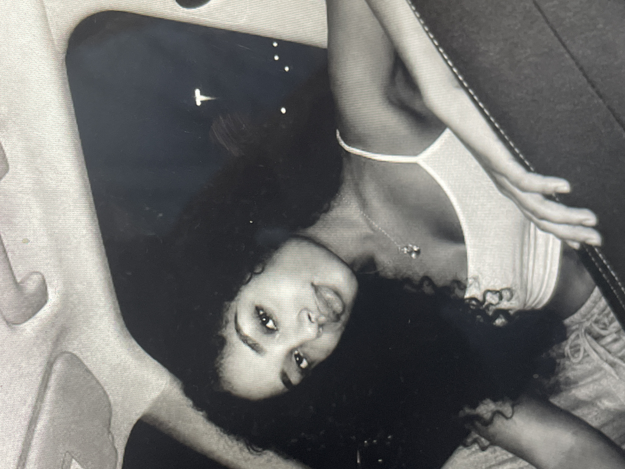 This black and white portrait features a young woman with long, dark, curly hair, captured in an indoor setting. She is positioned diagonally, appearing to be reclining or lying down, gazing directly at the camera with a relaxed and slightly playful expression, her tongue just visible between her lips. A small nose piercing is visible.
She is wearing a light-colored, possibly white or grey, strappy top and what appear to be patterned shorts or sweatpants. A necklace with a prominent pendant is visible around her neck. The background is mostly dark, with a distinct light-colored "T" shaped object and several small white dots, possibly reflections or light sources, in the upper left. Portions of light-colored, textured upholstery or furniture are visible in the foreground and along the right side of the frame. The overall image has a casual and engaging feel. No specific location cues within the image point to Sofia, Bulgaria, nor is any text visible. The time of day is indeterminate due to the indoor setting and lighting.