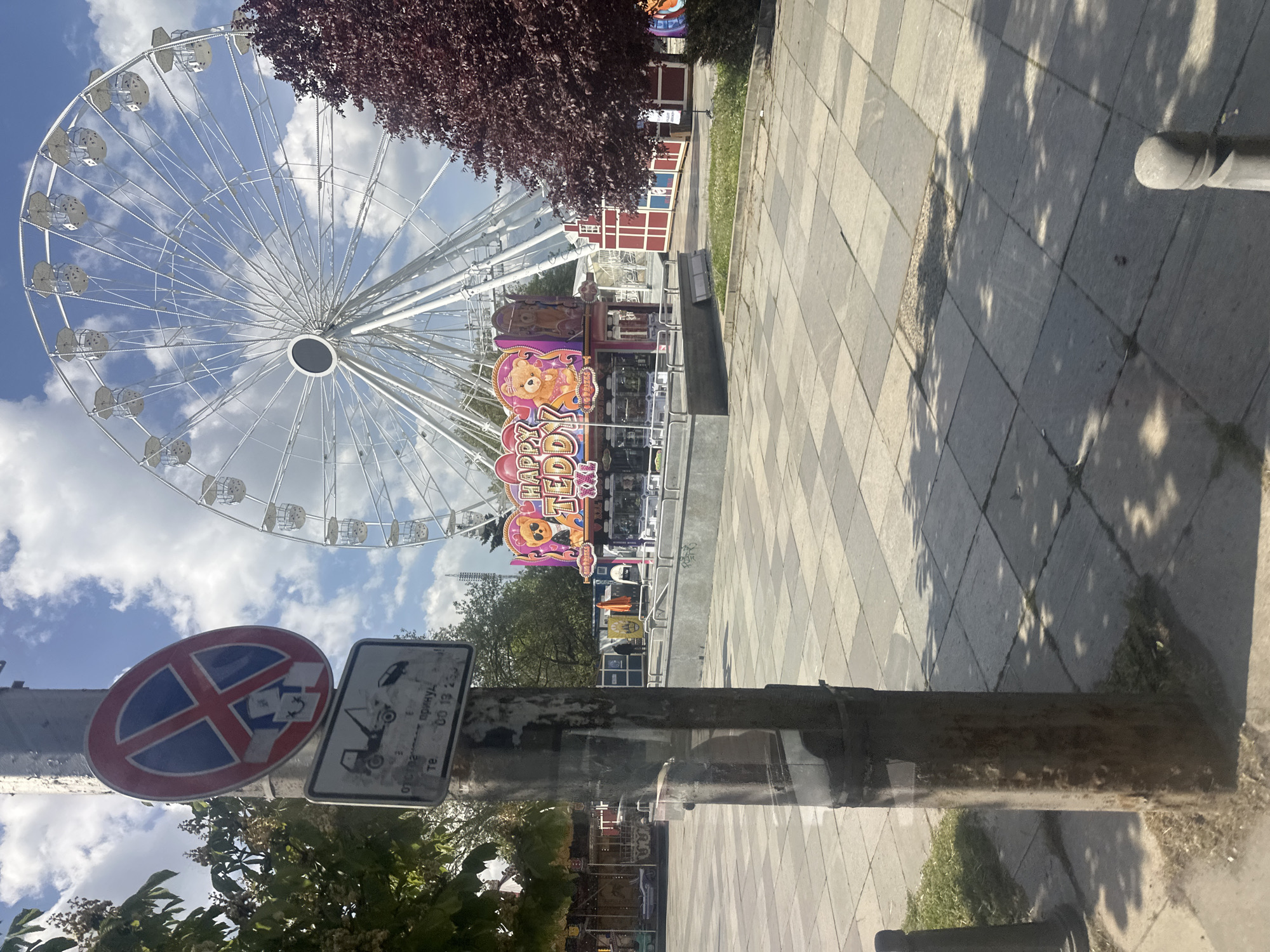 The image shows a view of a Ferris wheel at an amusement park in Sofia, Bulgaria. The Ferris wheel is white and large. A ride called "Happy Teddy" is visible. A red "no stopping" traffic sign and an information sign are attached to a pole in the foreground. Paved ground covers the area beside the amusement park. Trees and foliage are also present. The sky is blue with white clouds.