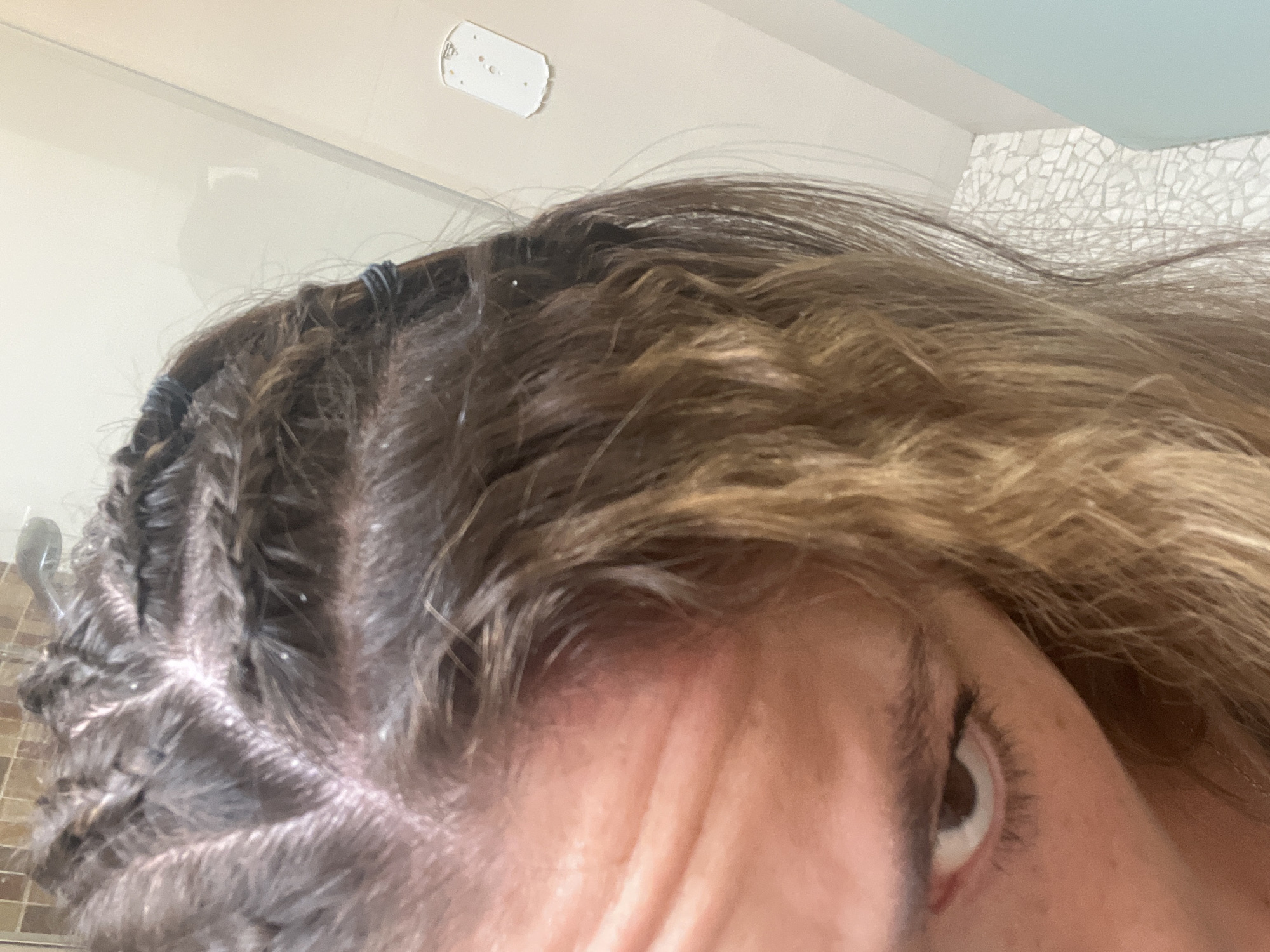 A close-up, high-angle shot shows a person's head and part of their face, including an eye. The person has braids close to the scalp on the left side of the image and loose, blondish-brown hair on the right. White flakes are visible on the scalp and in the braids, possibly dandruff. The background includes parts of a bathroom, with a white ceiling fixture, a section of decorative tiled wall, and a showerhead. There is no verifiable information in the image that places it in San Bernardino Tlaxcalancingo, Mexico.